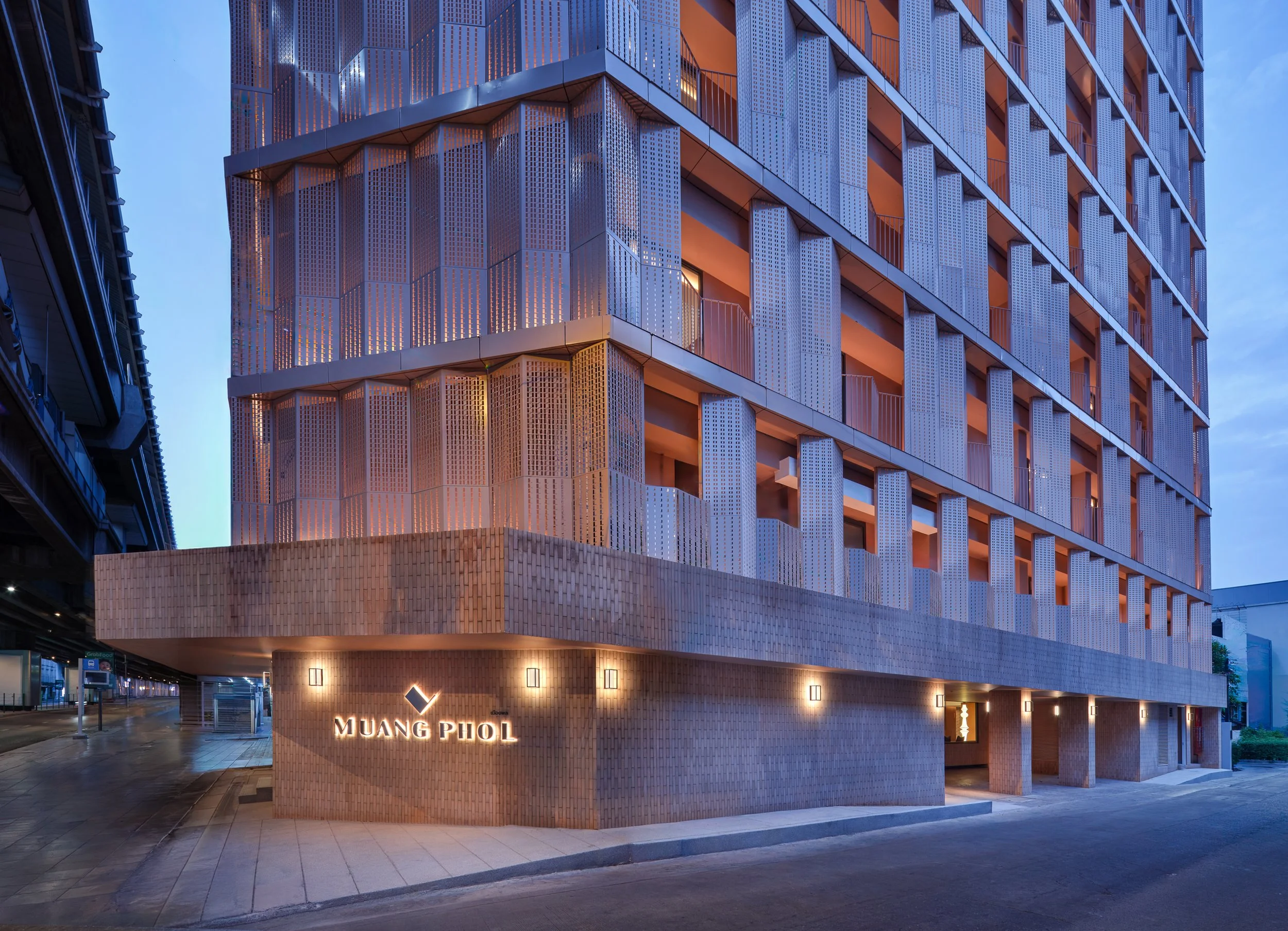 Nighttime view of a modern multi-story building with distinctive geometric and perforated metal facade, illuminated from within, with a sign that reads 'MUANG PHL' at the base.