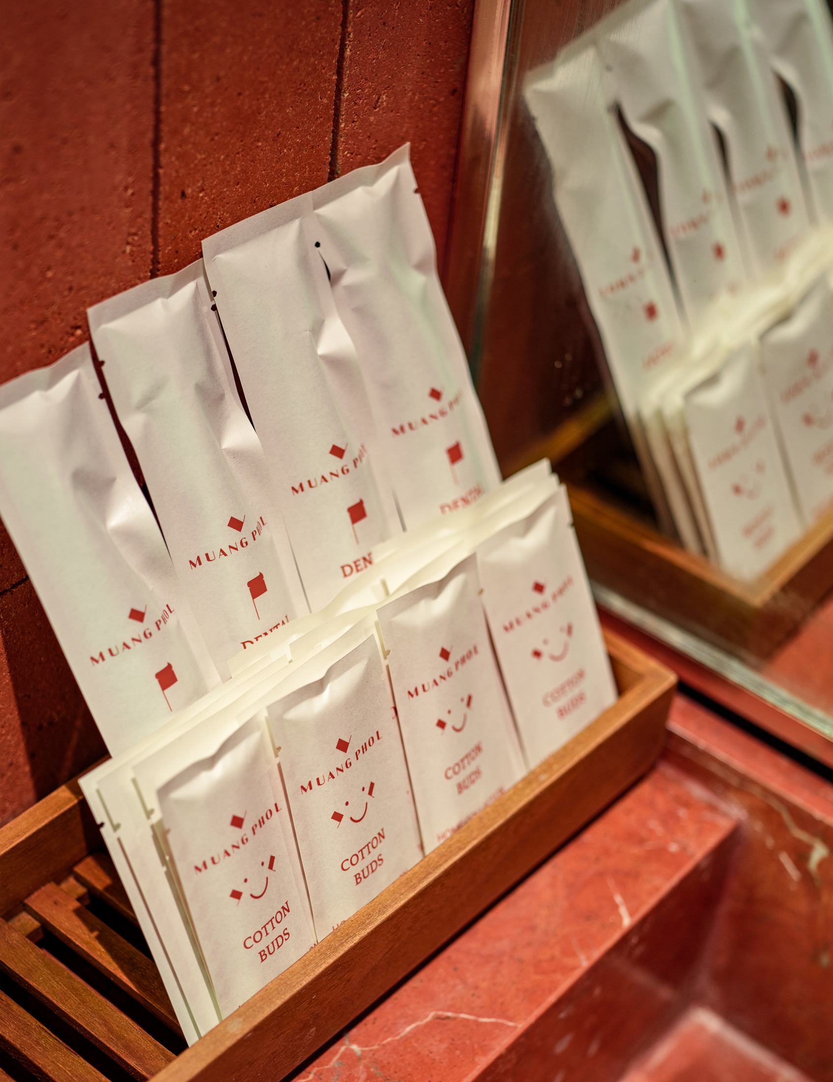 Display of small white paper packets labeled with red text and symbols, including 'Muang Phol', 'Cotton Buds', and a smiling face icon, arranged on a wooden surface.