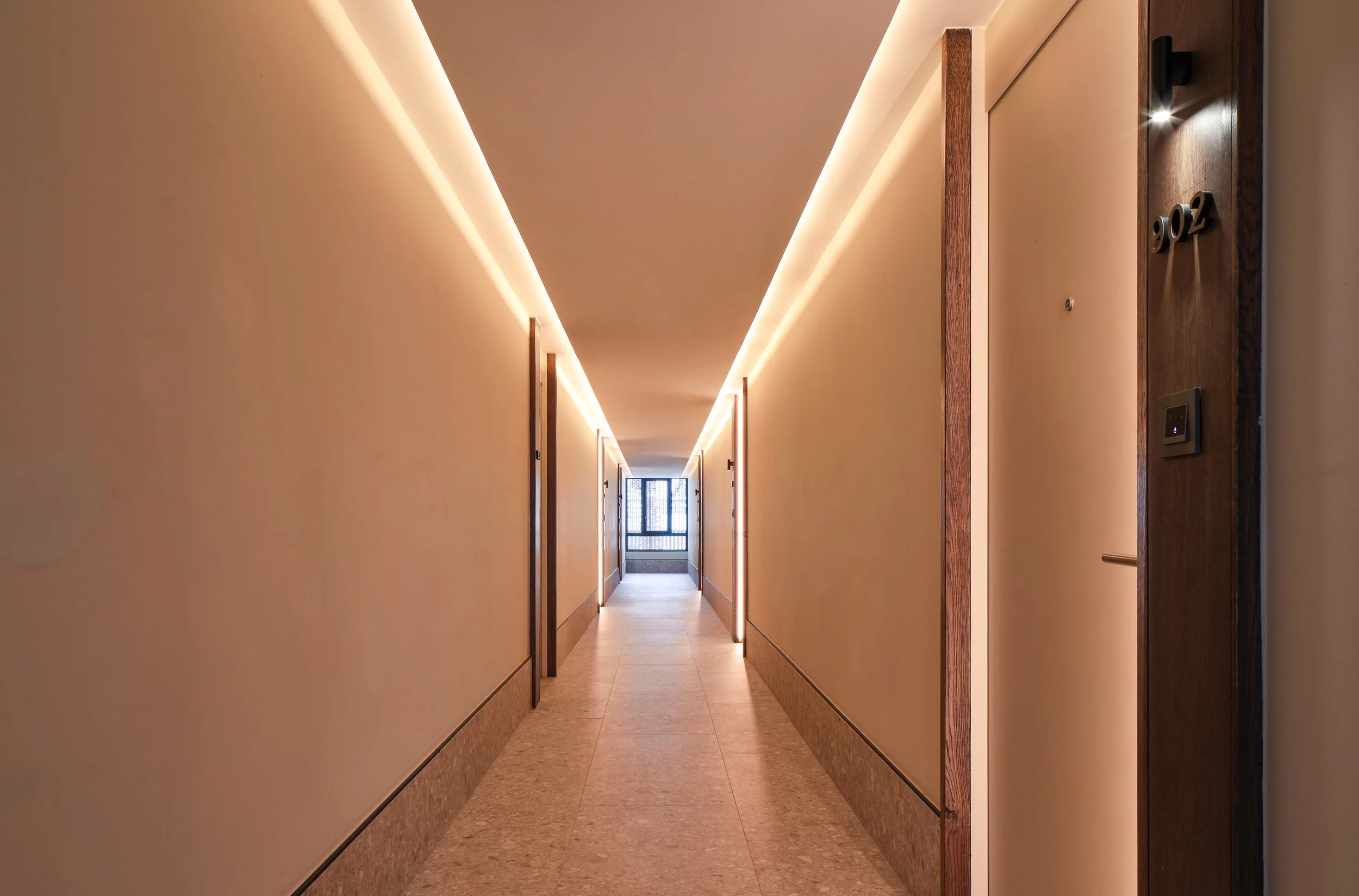 Long hotel corridor with beige walls, wood-framed doors, and warm lighting, leading to a glass door at the end.