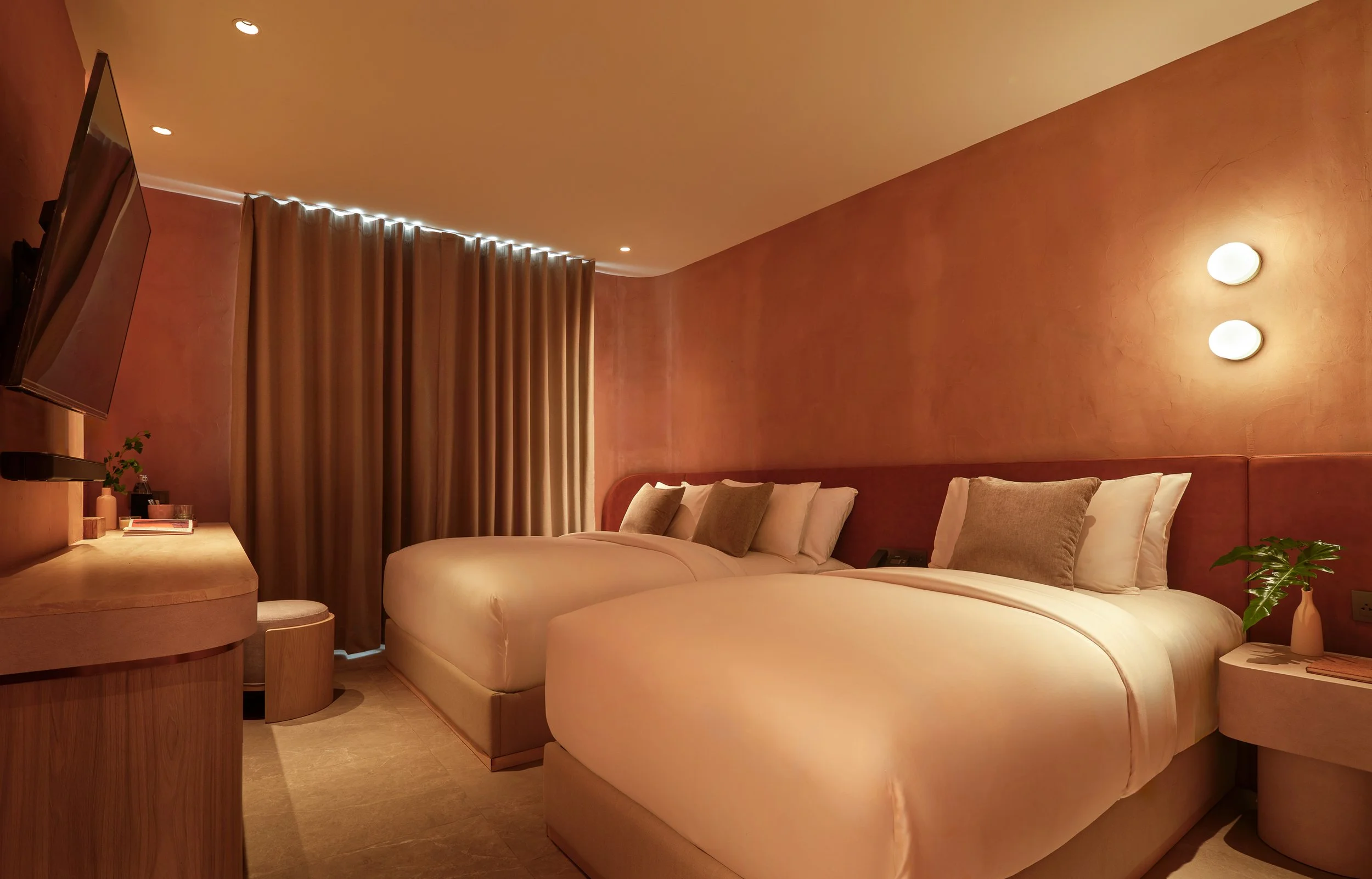 A cozy hotel room with a large bed, pink textured walls, soft lighting, beige curtains, a flat-screen TV, a desk with a small plant and decorative items.