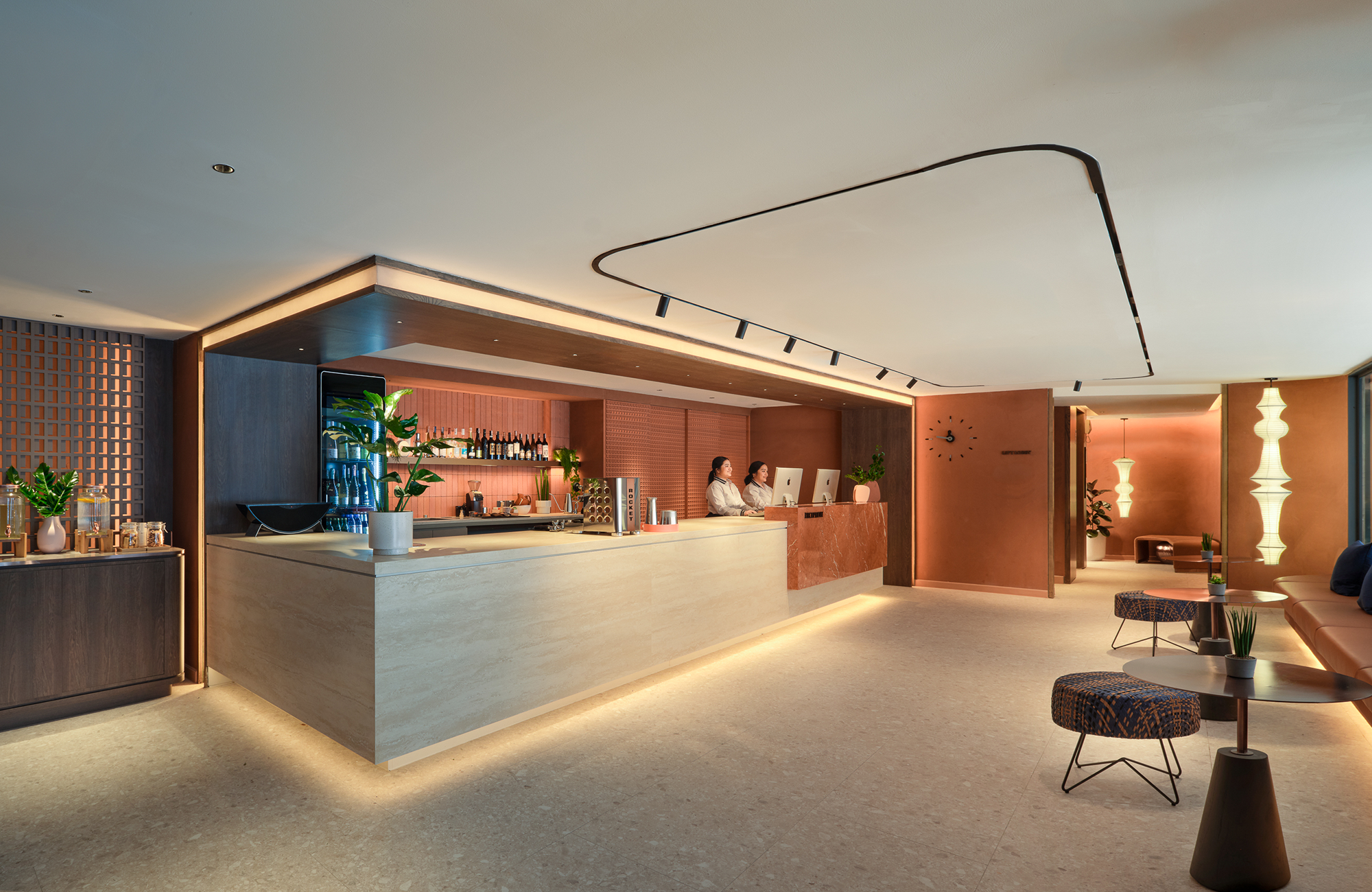 Modern hotel lobby with a reception desk, two receptionists working on computers, and seating area with patterned armchairs and small tables, warm lighting, and decorative plants.