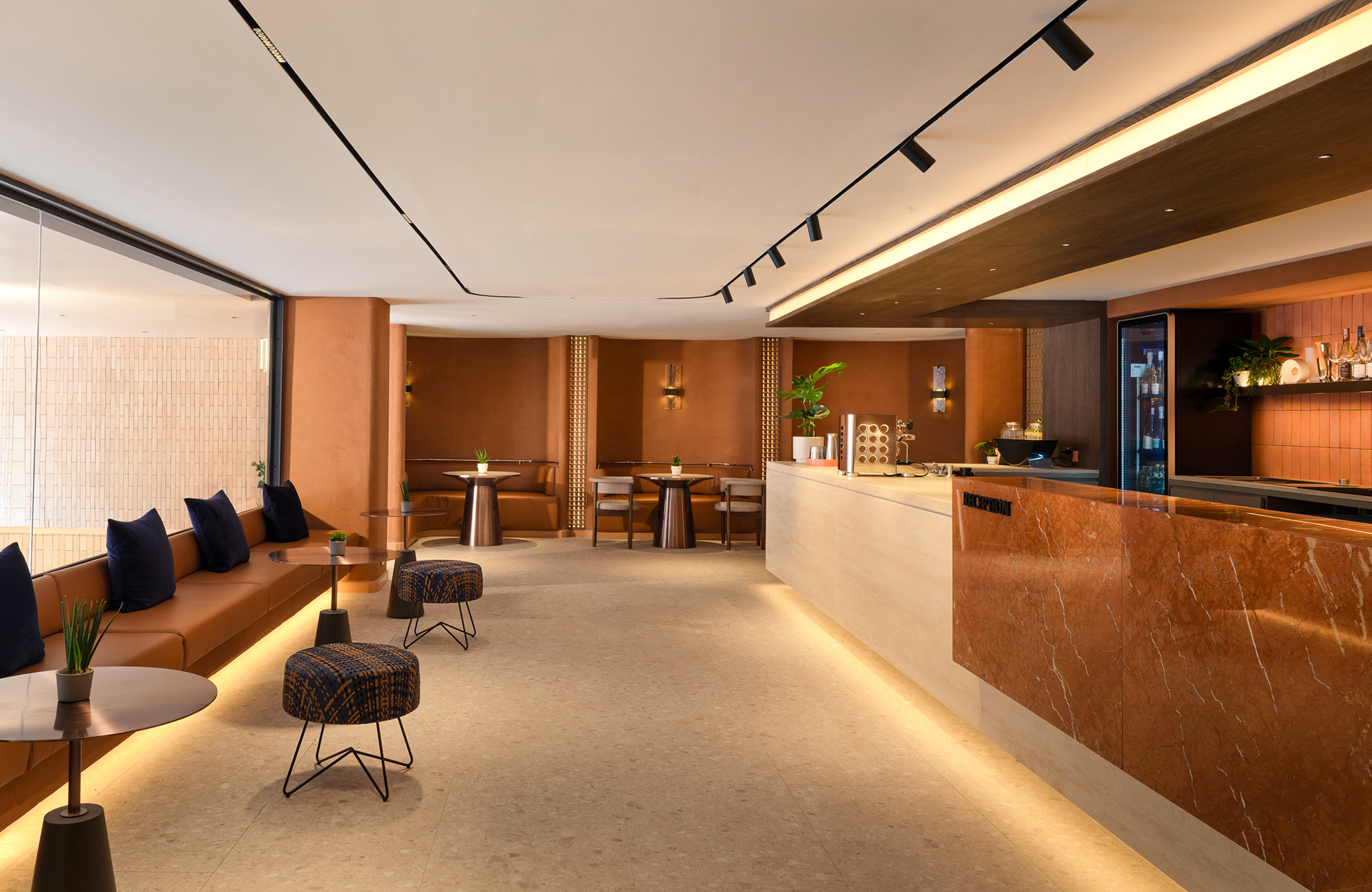 Modern hotel lobby with a Cushioned sofa, small tables with potted plants, a reception desk made of marble, and a bar area with shelves of bottles. The ceiling has contemporary track lighting, and the space features warm earth tones and contemporary decor.