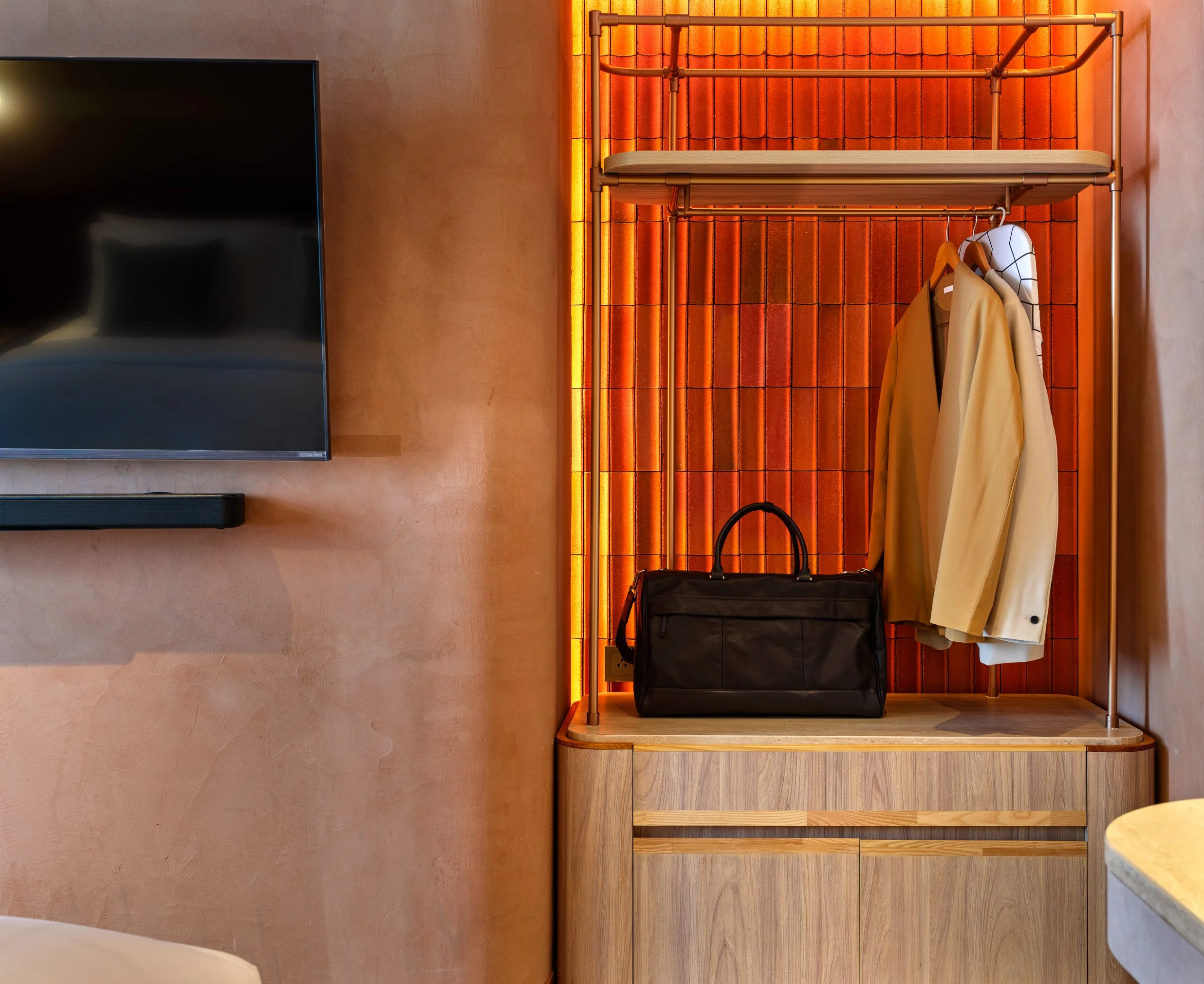 A wooden shelving unit with clothing and bags against an orange tiled wall with warm lighting.