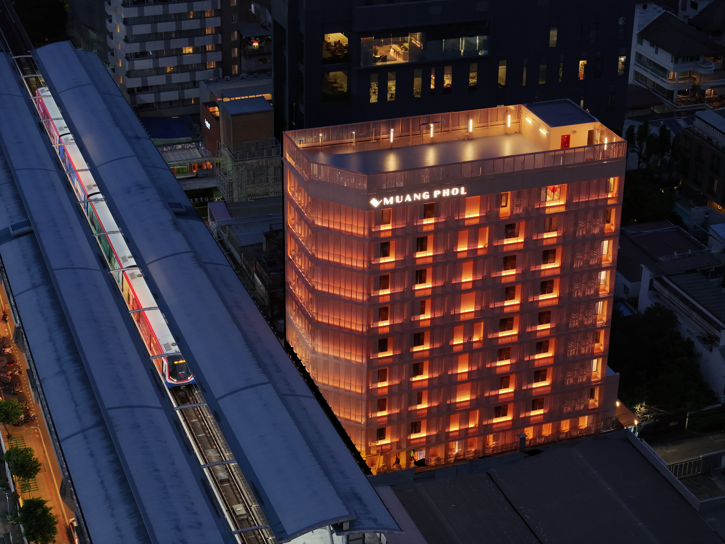 Night view of a tall, illuminated building with the sign 'Muang Phol,' and an elevated train passing by on the left side.