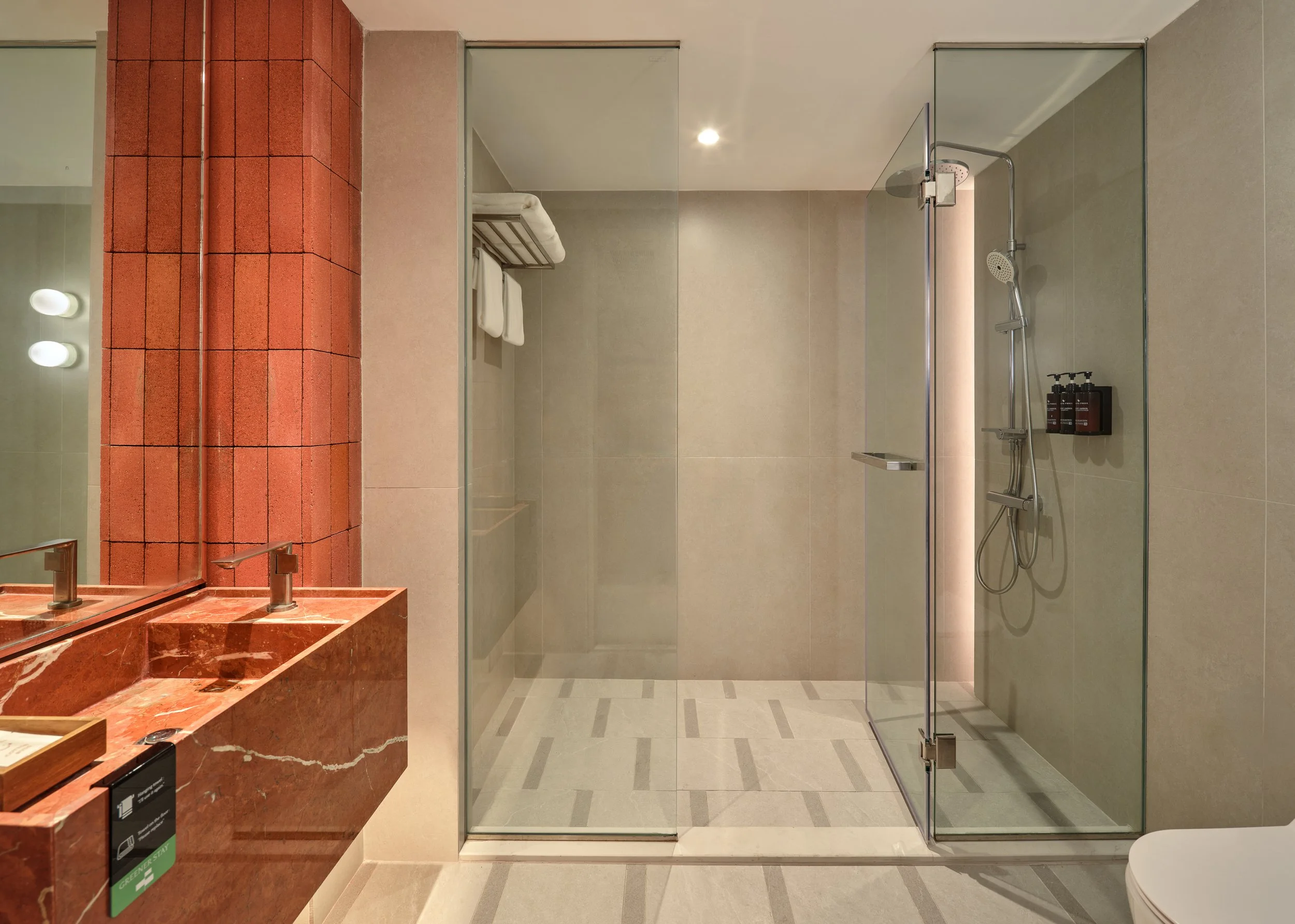 A modern bathroom featuring a glass-enclosed shower area with a showerhead and toiletries, a red marble vanity with sinks, a mirror, and a towel rack with white towels. The walls are tiled in neutral colors, and there is a ceiling light fixture.