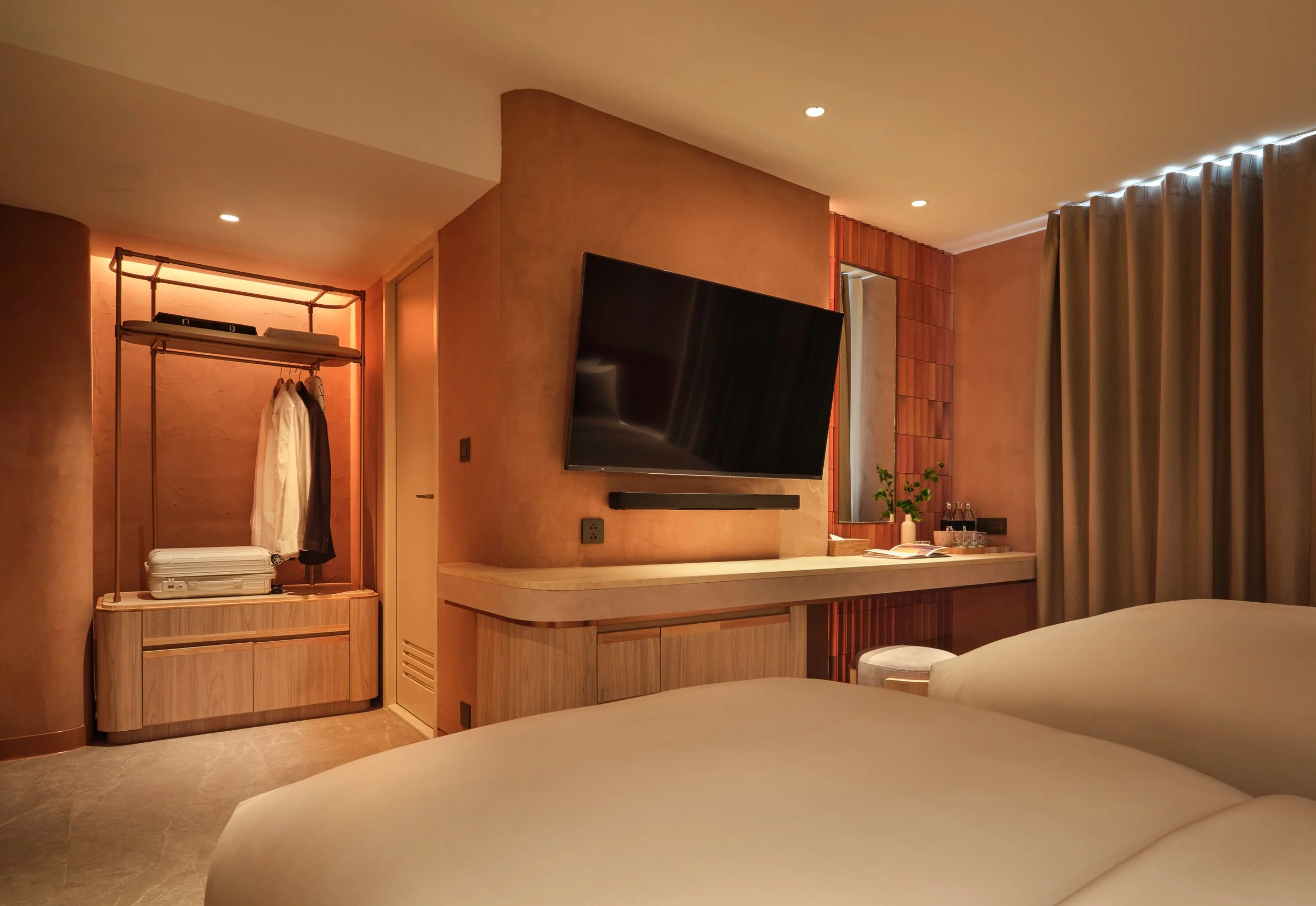A cozy hotel room or suite with a flat-screen TV, a mirror, a bed partially visible in the foreground, a wooden wardrobe with hanging clothes, and a curtain covering a window or balcony, with warm lighting and modern decor.