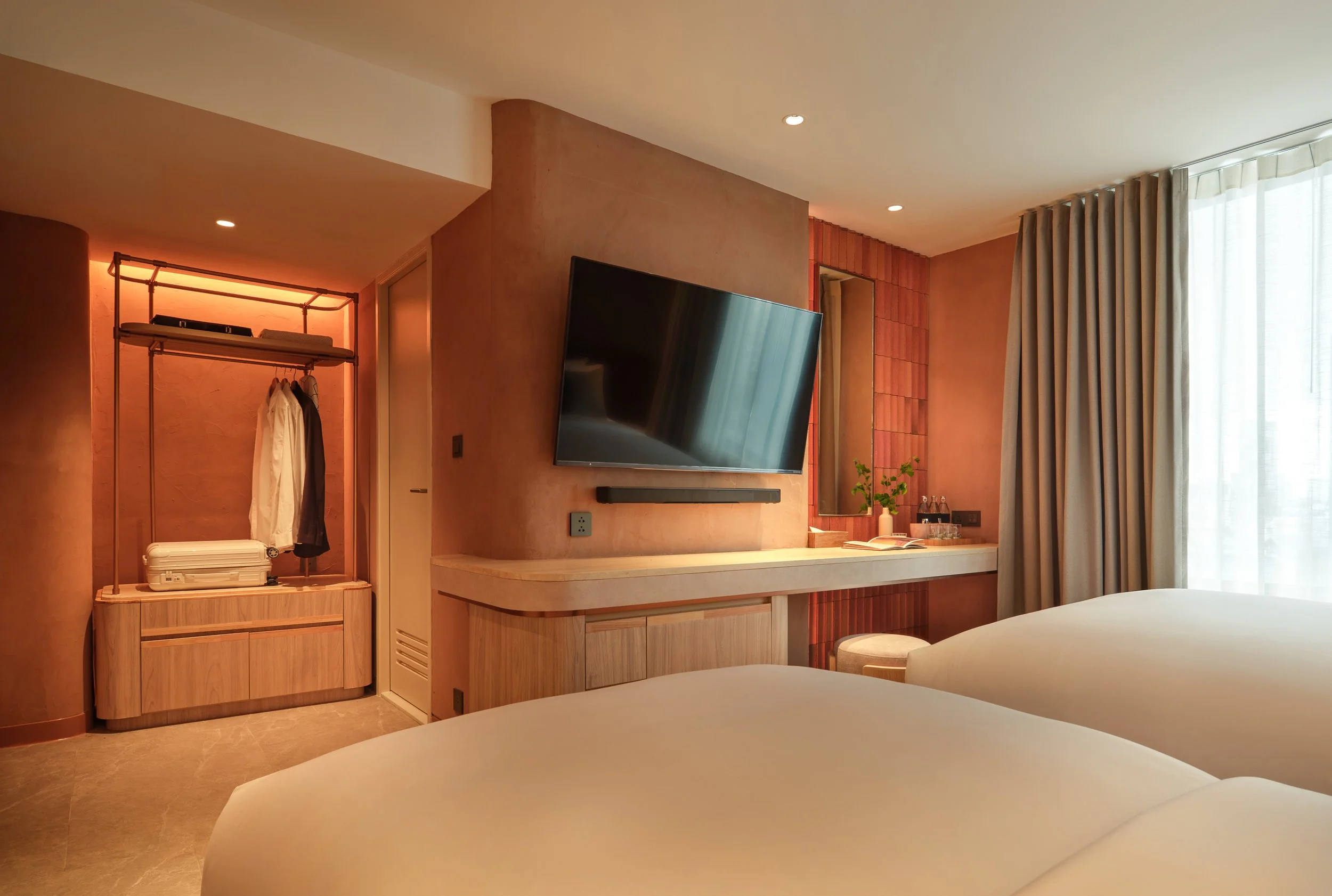 Modern hotel room with a large flat-screen TV mounted on a beige wall, a wooden desk with decorative plants, beige curtains covering a window, and a seating area with beds in the foreground.