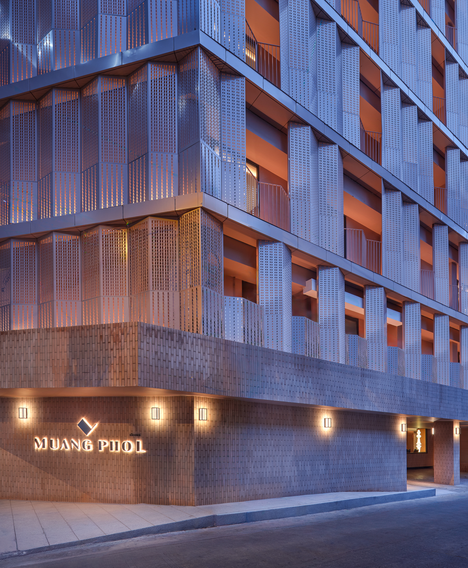 Exterior view of a modern building with a distinctive perforated metal facade, illuminated at night, with a sign reading 'Muang Phol' and a geometric logo at the entrance.
