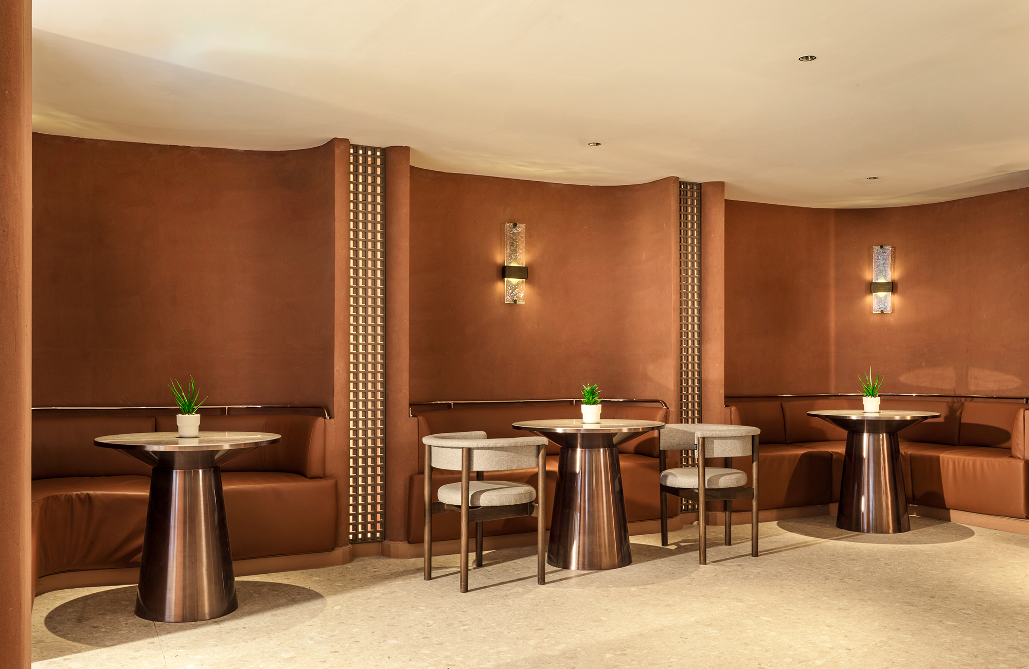A cozy hotel lobby or lounge with brown upholstered seating arranged along a curved wall, small round tables with metallic bases, and potted green plants as decor. The wall has vertical decorative panels and is lit by wall sconces.