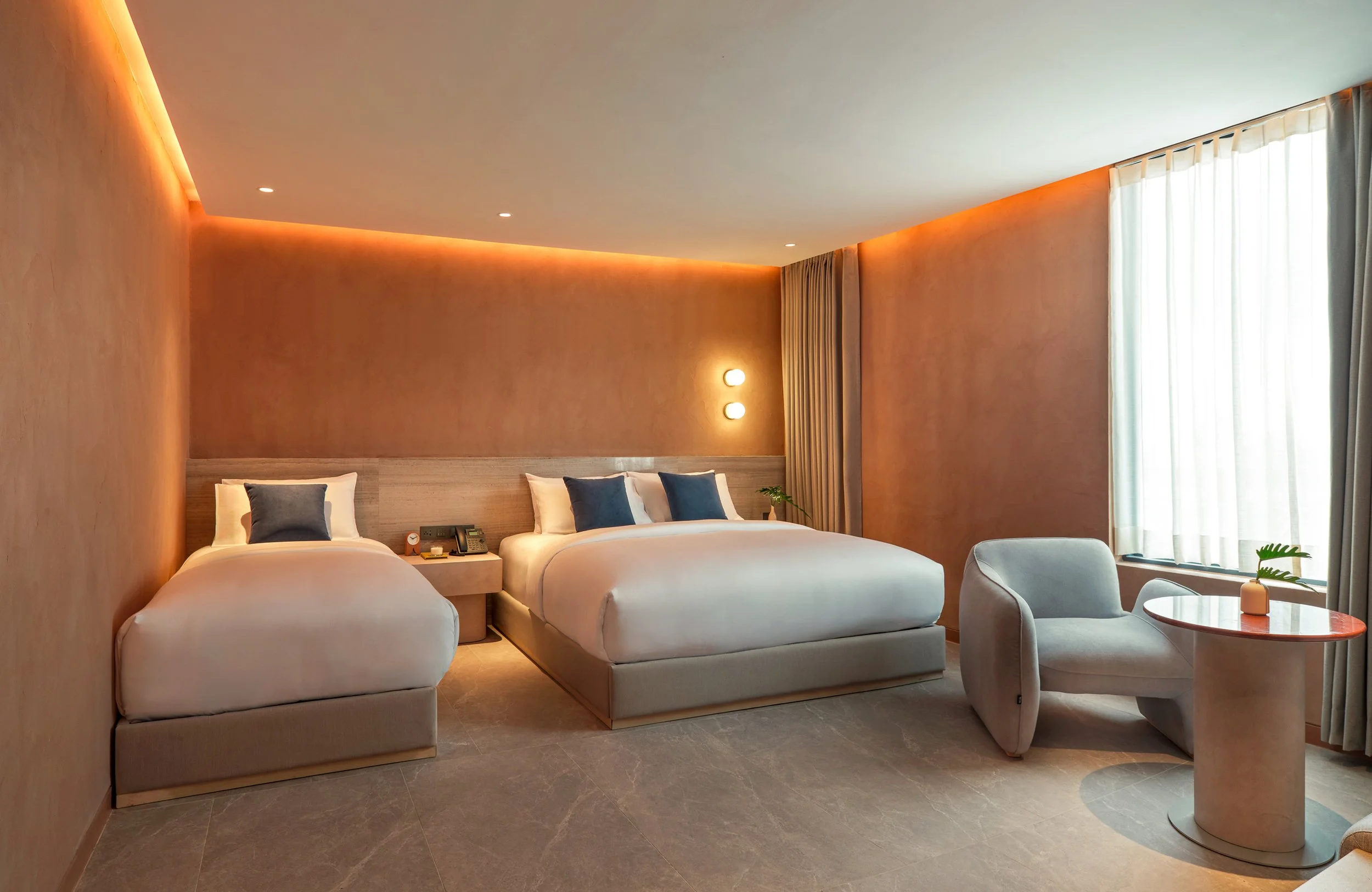 A modern hotel room with two beds, a sitting area, and large window with curtains letting in natural light.