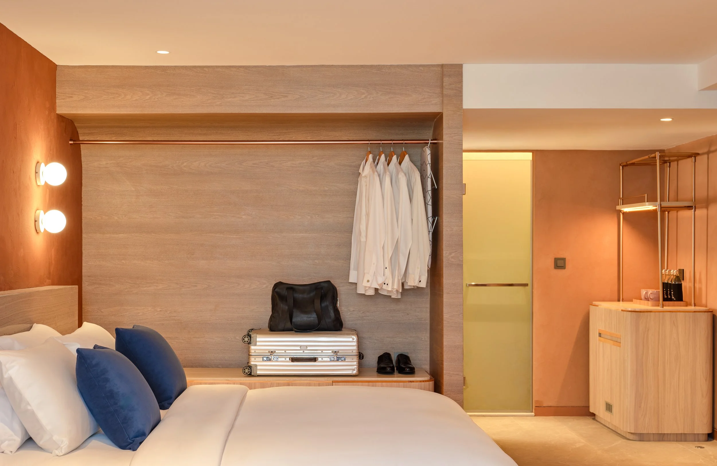 Hotel room with bed, navy pillows, built-in hanging closet with white shirts, black bag, suitcase, shoes, bedside table, and decorative lighting.