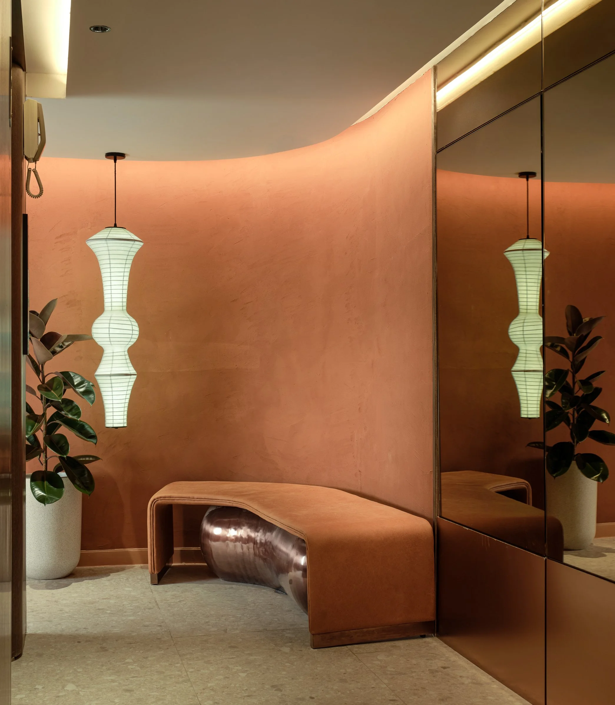 Interior space with a curved pink wall, a wooden bench, a tall beige planter with a large leafy plant, a mirrored wall reflecting a paper lantern hanging from the ceiling, and a beige floor.