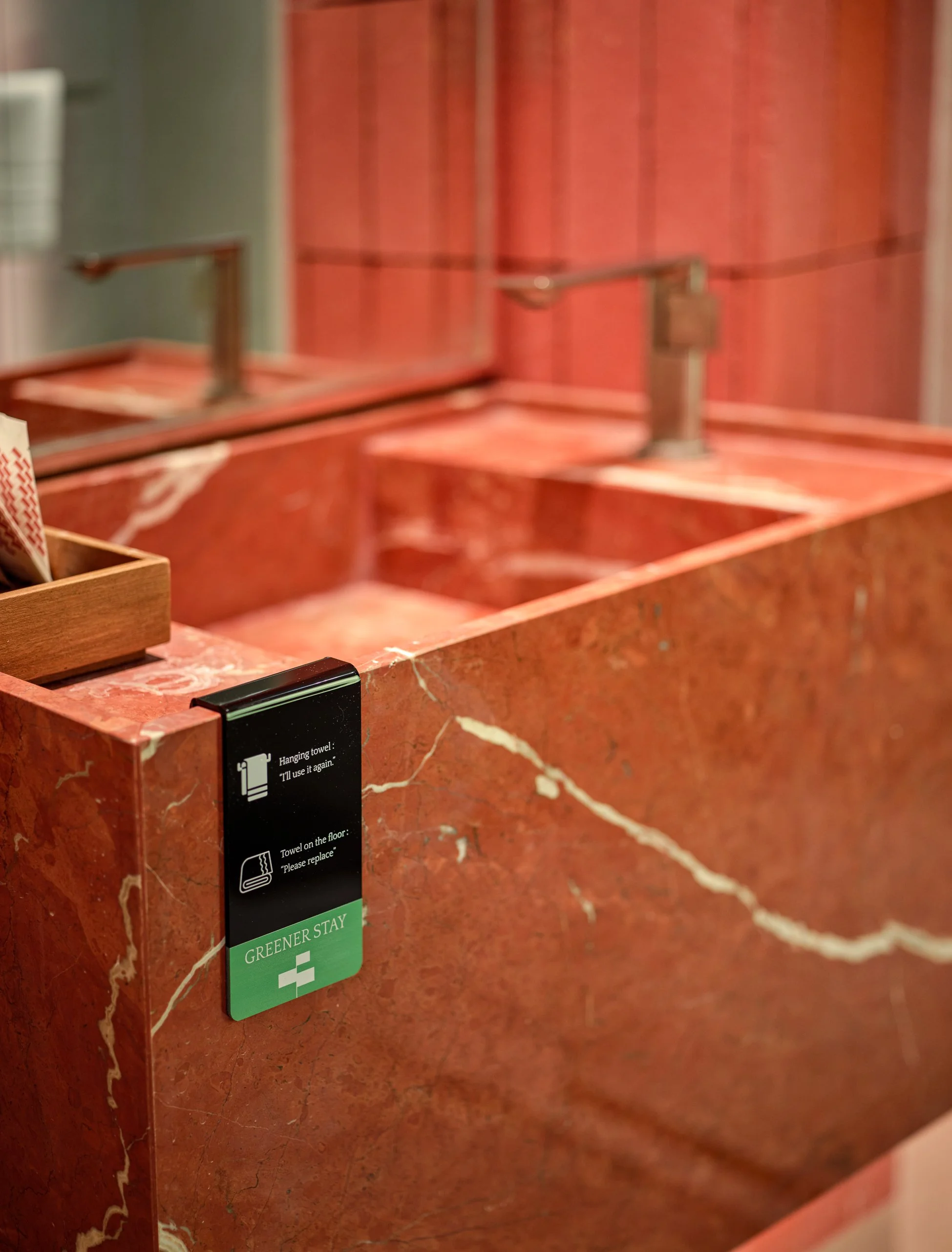 A red marble countertop with a sign that says 'Hanging towel: 'I'll use it again''. The countertop has a black soap dispenser, and there are handrails and a mirror in the background.
