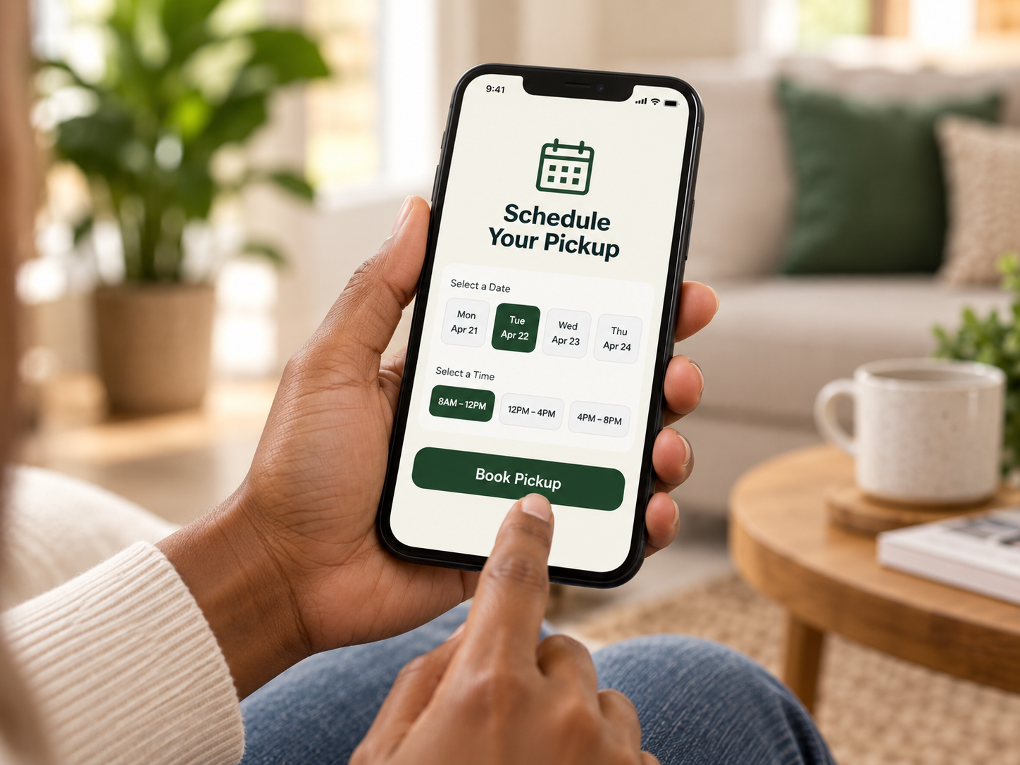 Person holding a smartphone with a pickup scheduling app open, showing options to select a date and time for pickup, sitting in a cozy living room with plants and a coffee mug on a wooden table.