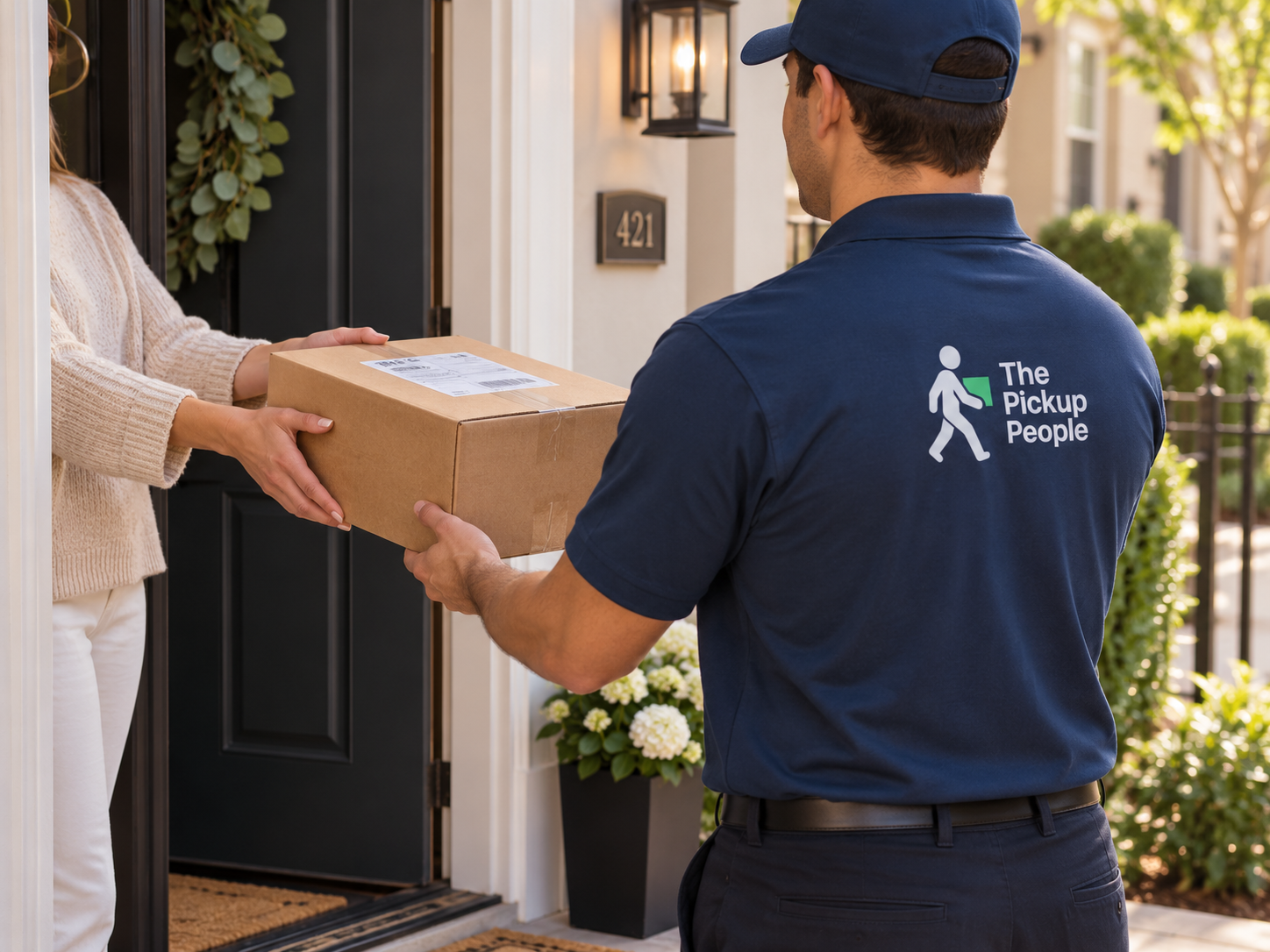 A package delivery person from The Pickup People handing a cardboard box to a woman at her doorstep on a sunny day.