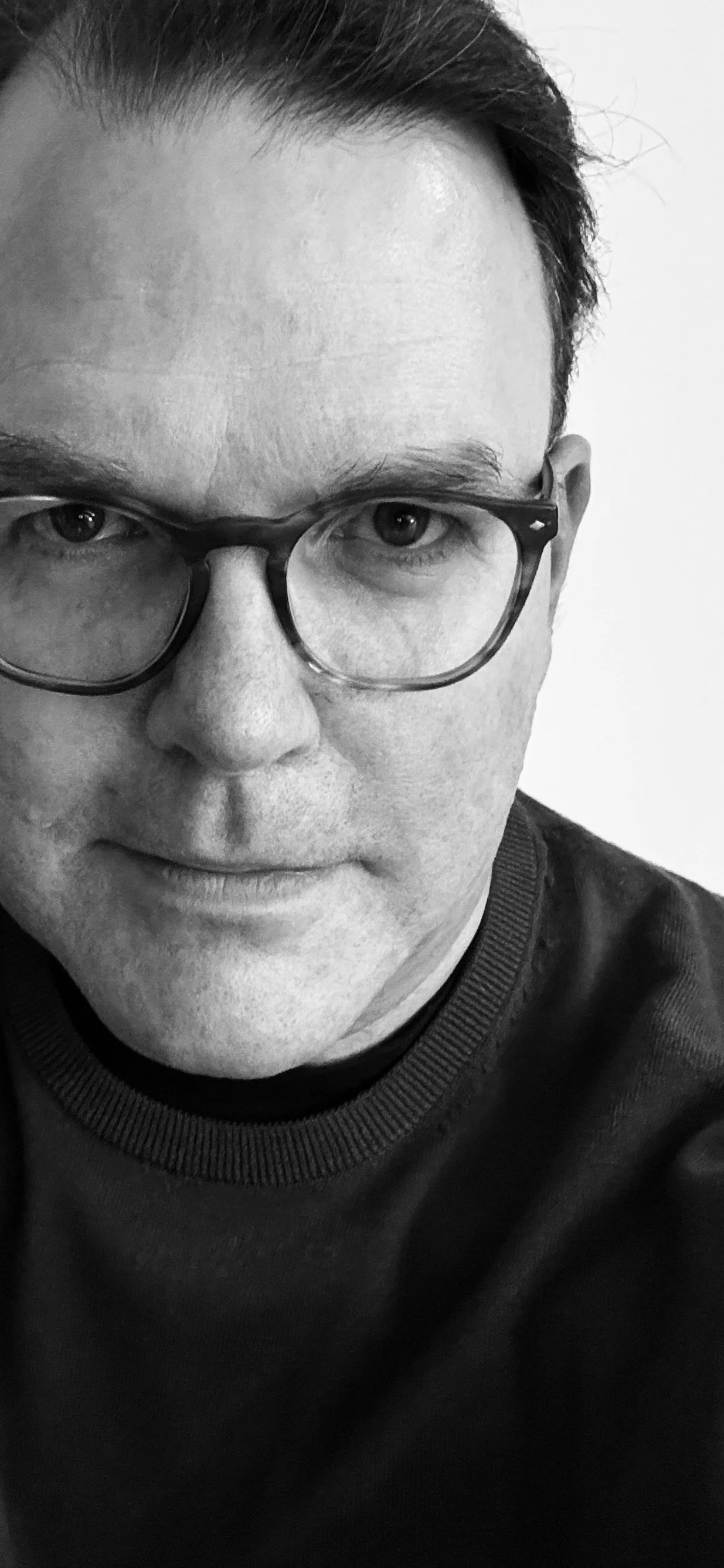 Black and white close-up portrait of a man wearing glasses and a dark shirt, looking into the camera with a neutral expression.