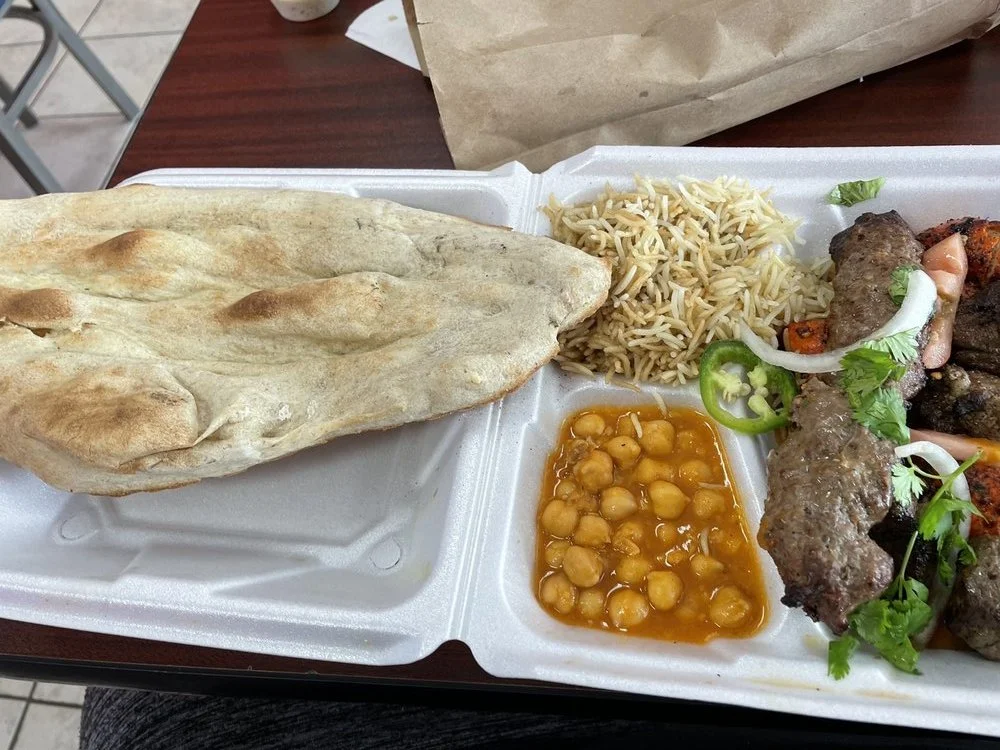 A styrofoam plate with naan bread, rice, chickpeas in sauce, and skewered grilled kebabs garnished with herbs and vegetables.