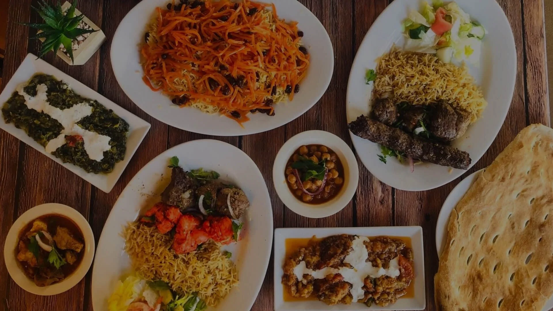Assorted Indian dishes on white plates, including rice, grilled meat, vegetable curries, flatbread, and lentil salads, arranged on a wooden table.