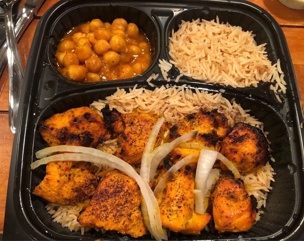 A meal with grilled chicken, basmati rice, chickpea curry, and cooked chickpeas in sauce, served in a black compartmental tray.