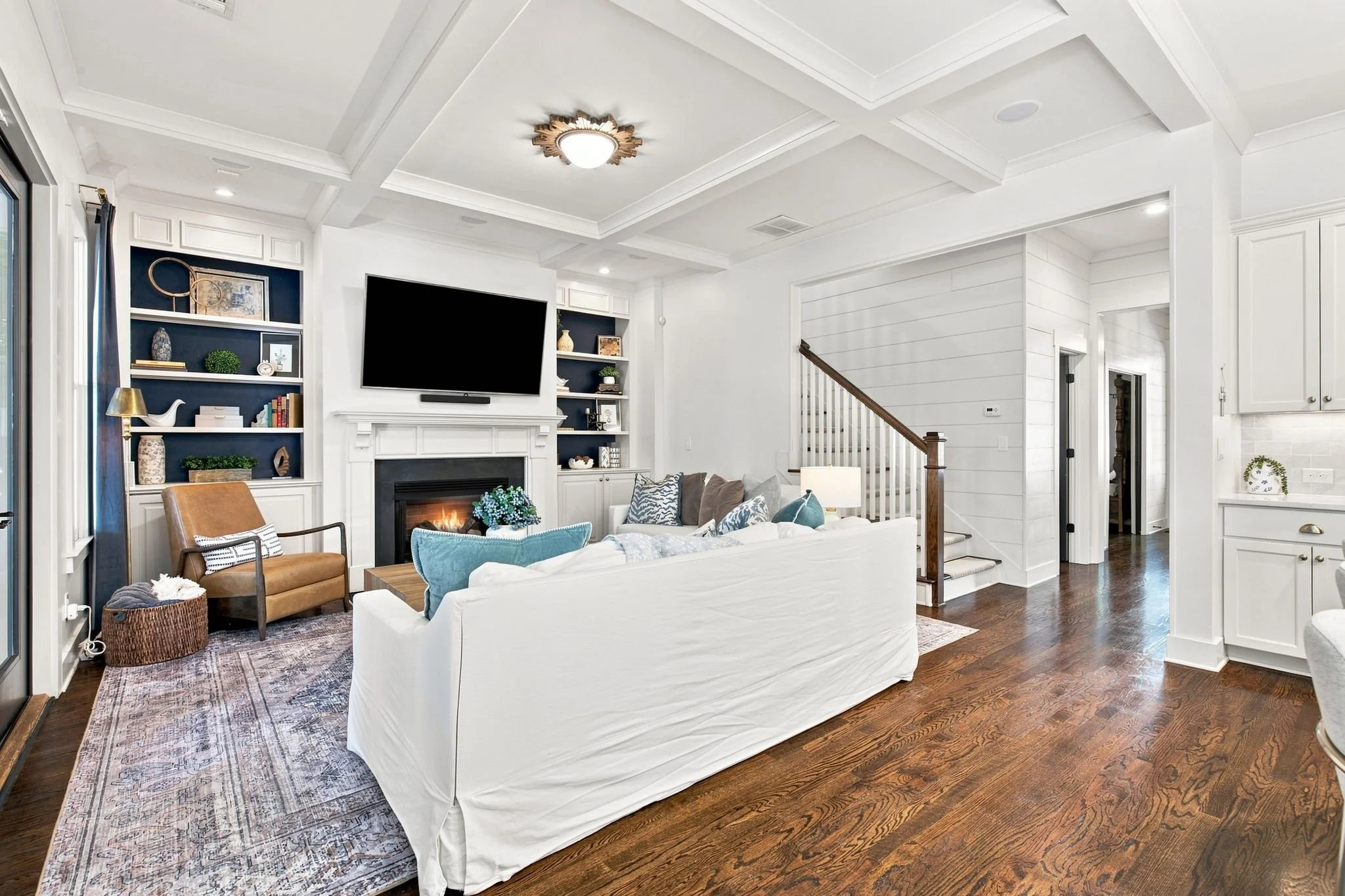 Bright living room with white walls, a white sofa, a brown armchair, a flat-screen TV above a fireplace, bookshelves with decor, hardwood floors, and a ceiling with recessed lighting and a decorative light fixture.