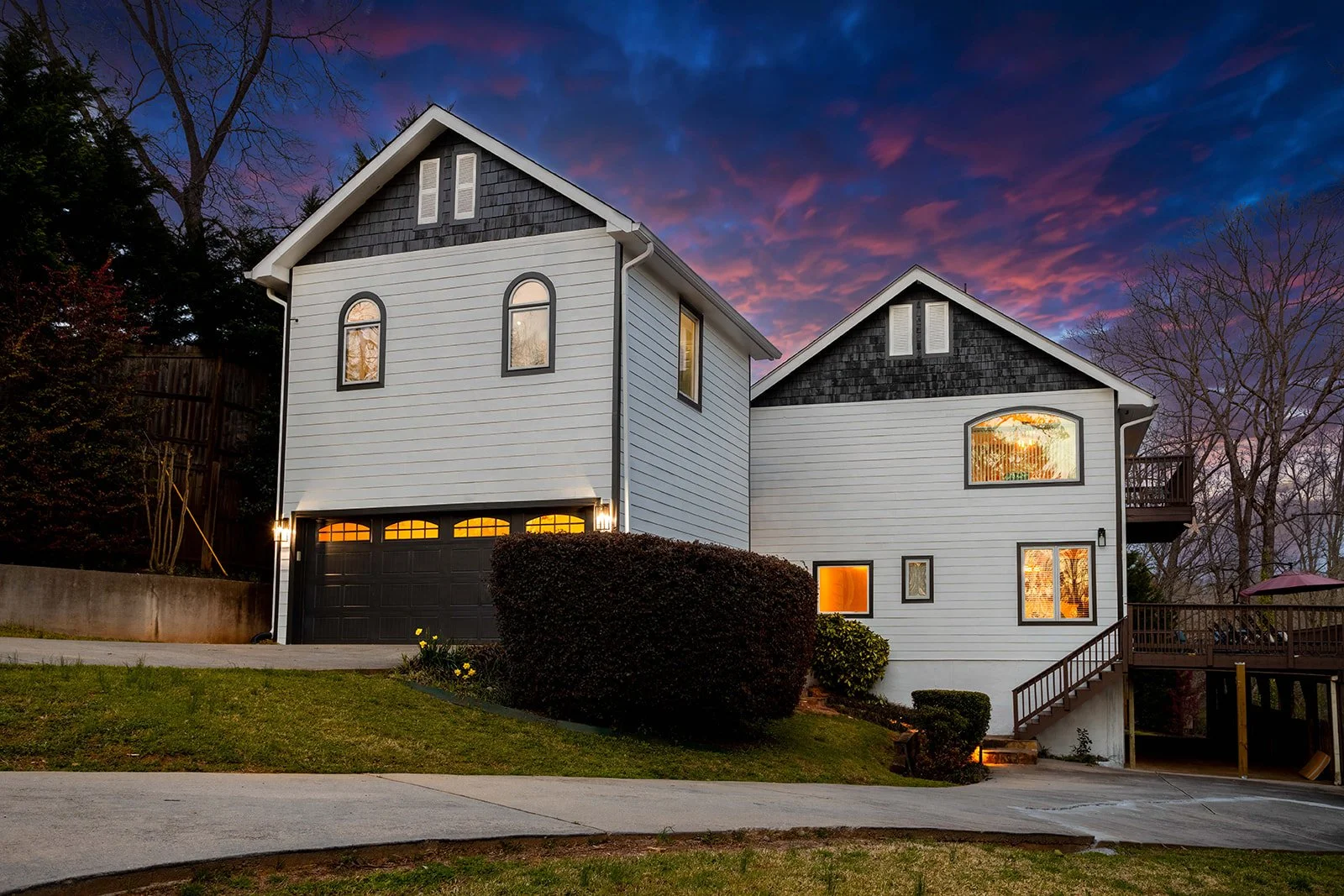 Two-story white house with black accents and a dark garage door, set against a vibrant sunset sky with purple and pink clouds. The house features multiple windows, some illuminated from within, and a front yard with bushes and a concrete sidewalk.