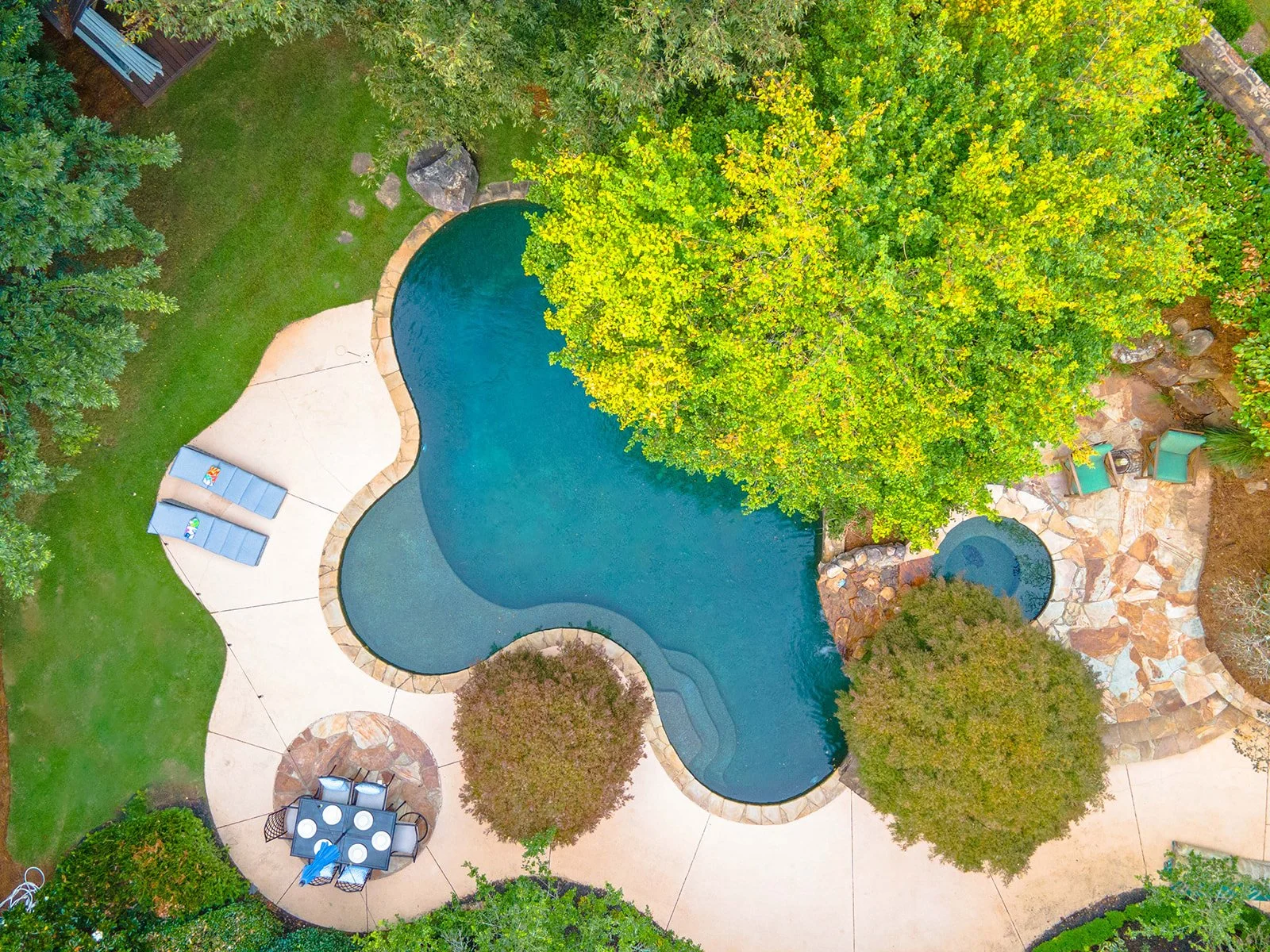 A backyard with a curvy swimming pool surrounded by green grass, trees, and a stone patio. There are two lounge chairs with towels on a concrete area near the pool. Additionally, there is a patio table with chairs, a small round hot tub, and various 