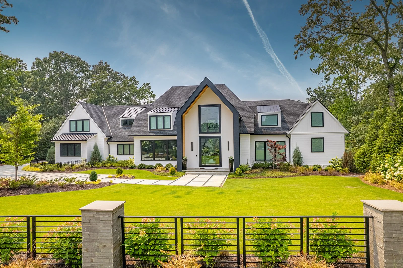 Modern white house with black trim and large windows, surrounded by a manicured lawn, trees, and a black fence, under a partly cloudy sky.