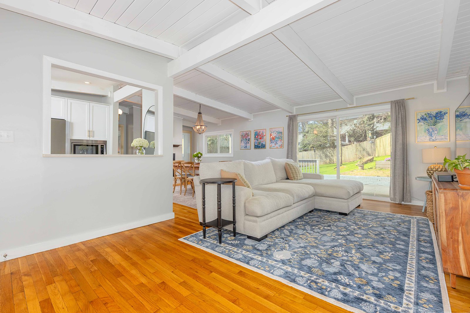 Living room with white sectional sofa, blue patterned rug, wood flooring, and large sliding glass door overlooking backyard with grass and trees.
