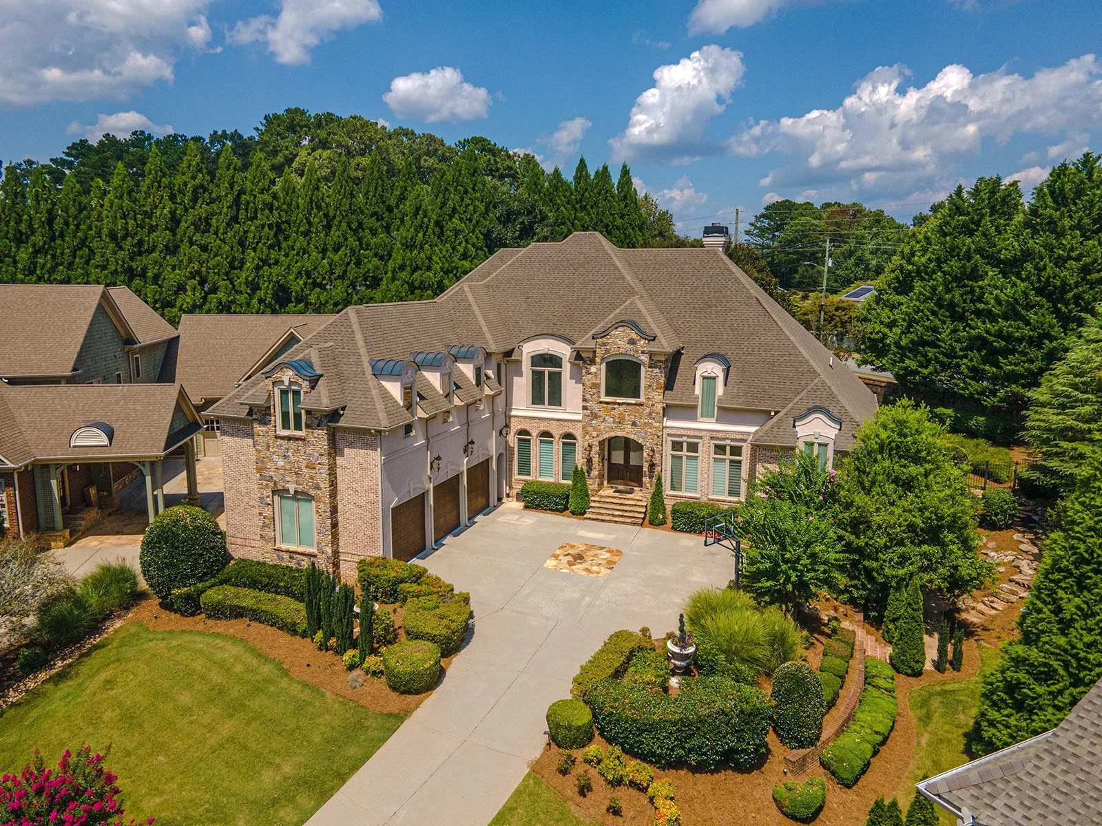 A large two-story house with a stone facade, multiple windows, and a gated driveway surrounded by lush greenery and trees