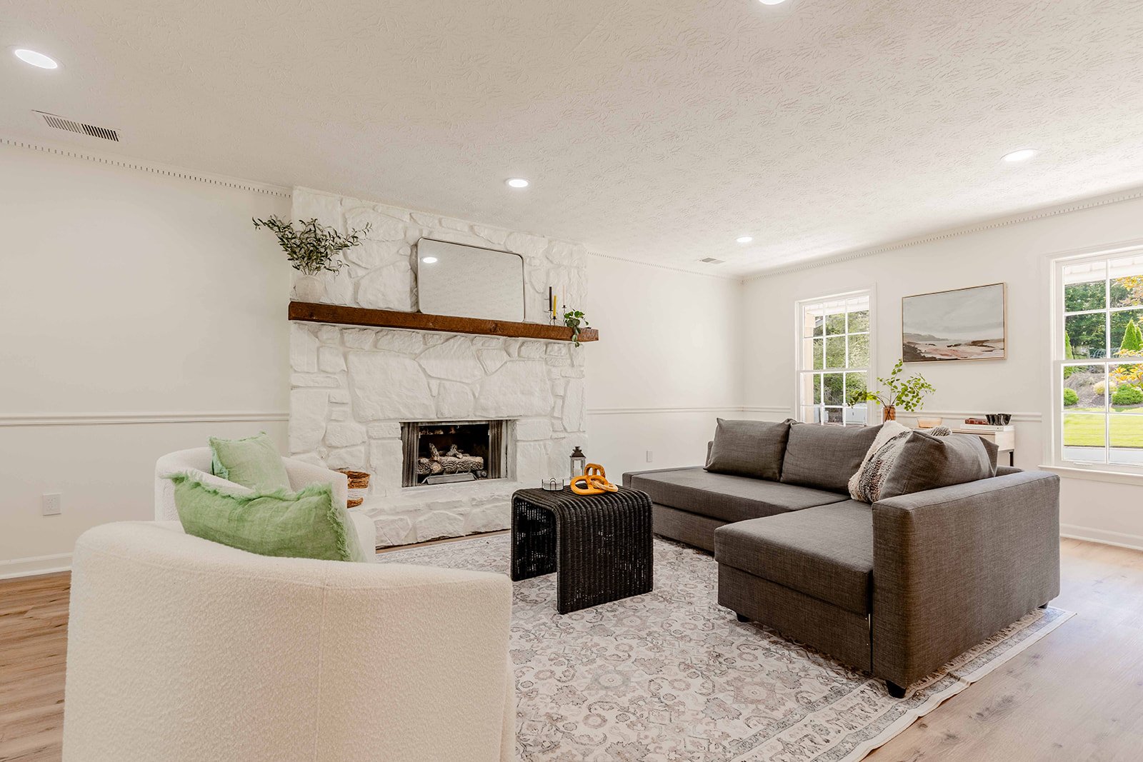 Living room with a white stone fireplace, gray sectional sofa, white armchair with green pillow, patterned rug, and large windows with greenery outside