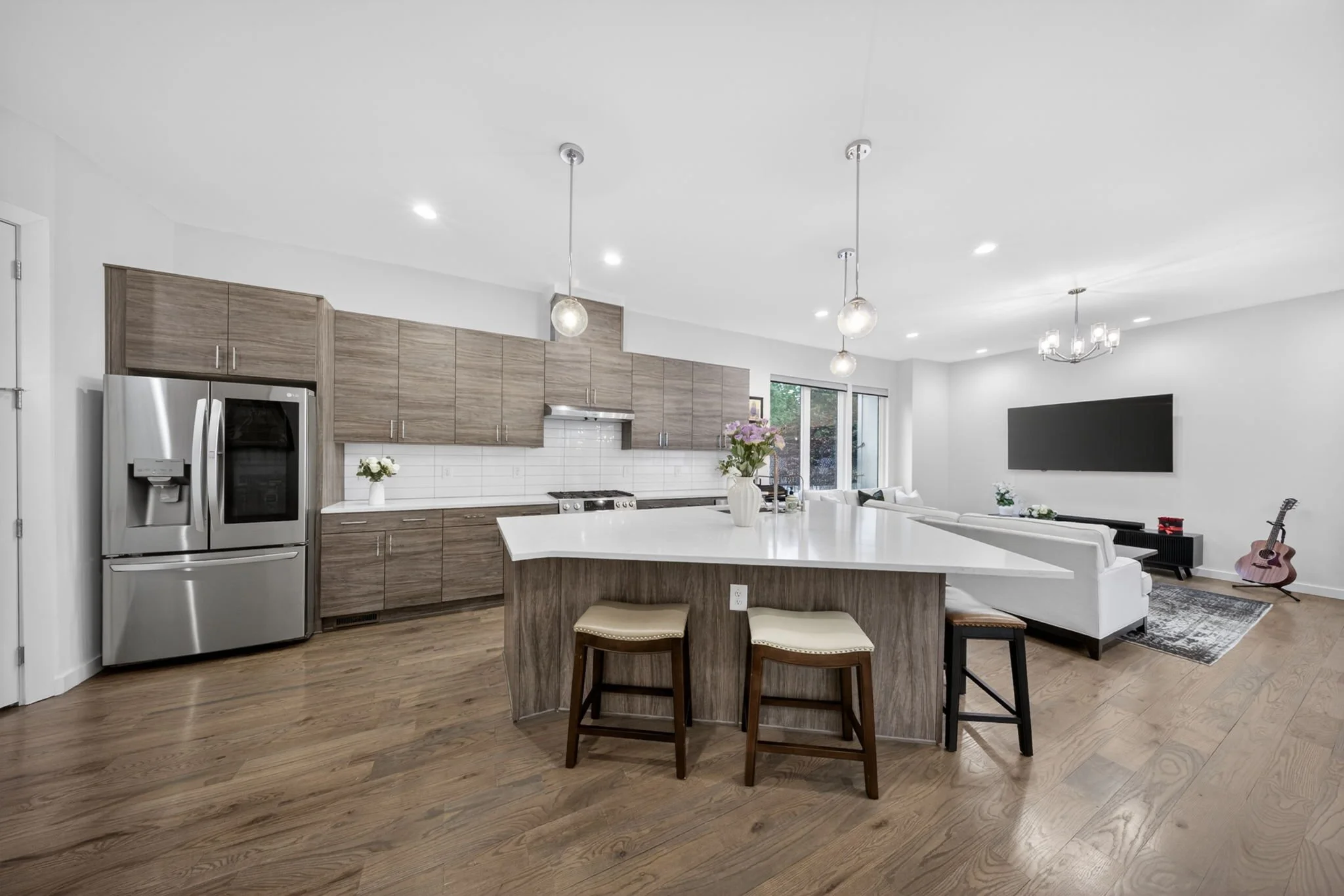 Modern open-plan kitchen and living area with wood cabinets, stainless steel refrigerator, white countertop island, dining chairs, white sofa, TV mounted on wall, guitar, and large windows.