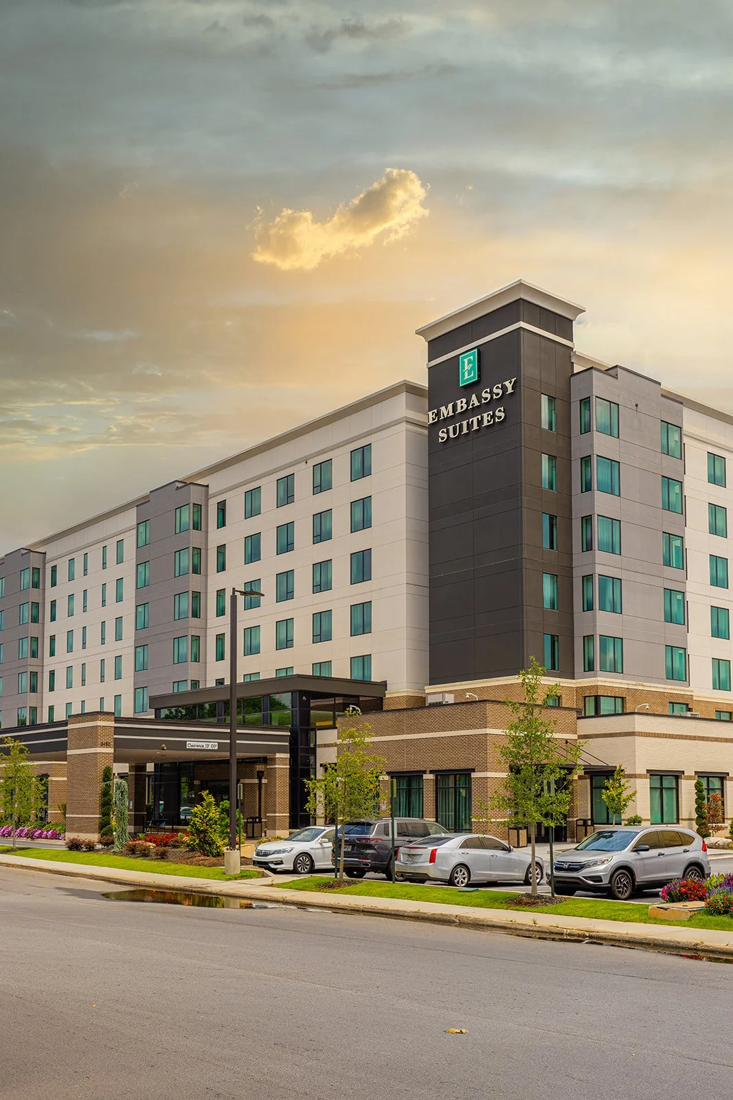 Exterior of a modern multi-story hotel named Embassy Suites with parked cars in front and small trees along the sidewalk, under a partly cloudy sky.