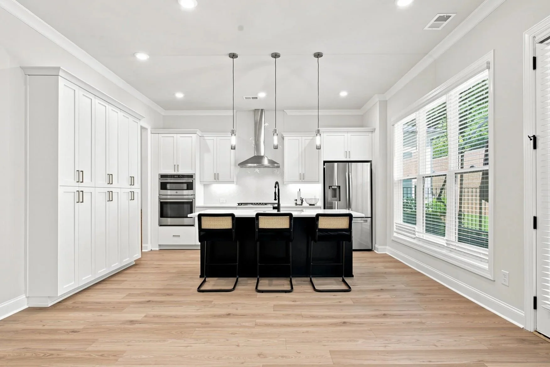 Bright, modern kitchen with white cabinets, black island with seating, stainless steel appliances, large windows with blinds, and hardwood floors.