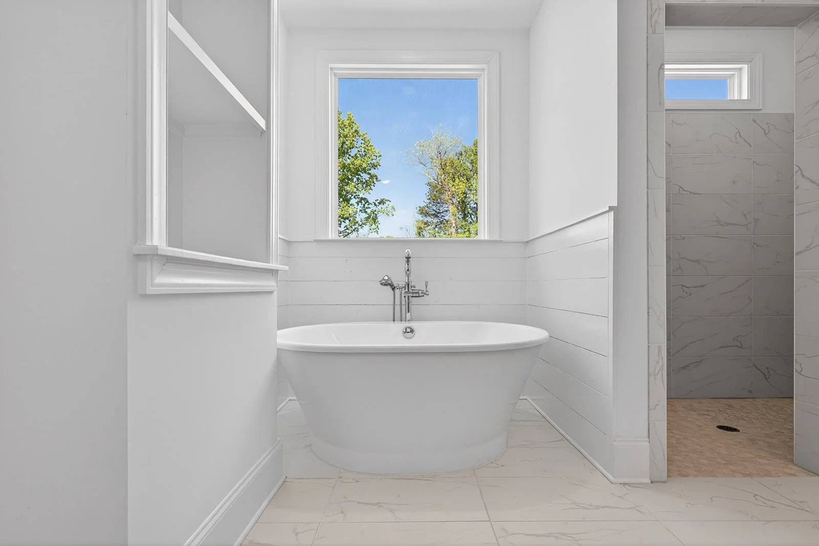 White bathtub near a window with a view of trees and blue sky, in a modern bathroom with white walls and marble tile accents.