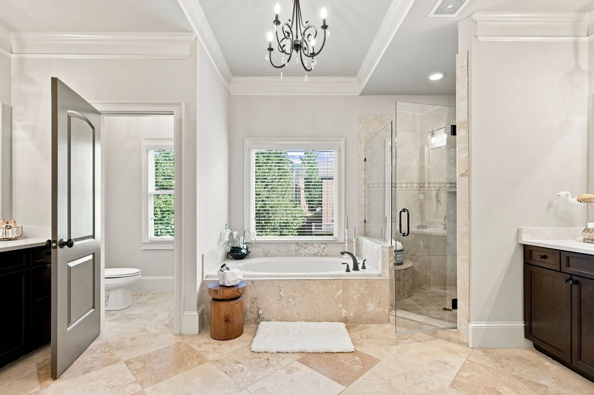A bright and spacious bathroom with a chandelier hanging from the ceiling, a bathtub beneath a large window, a glass-enclosed shower, and dark wooden cabinets.