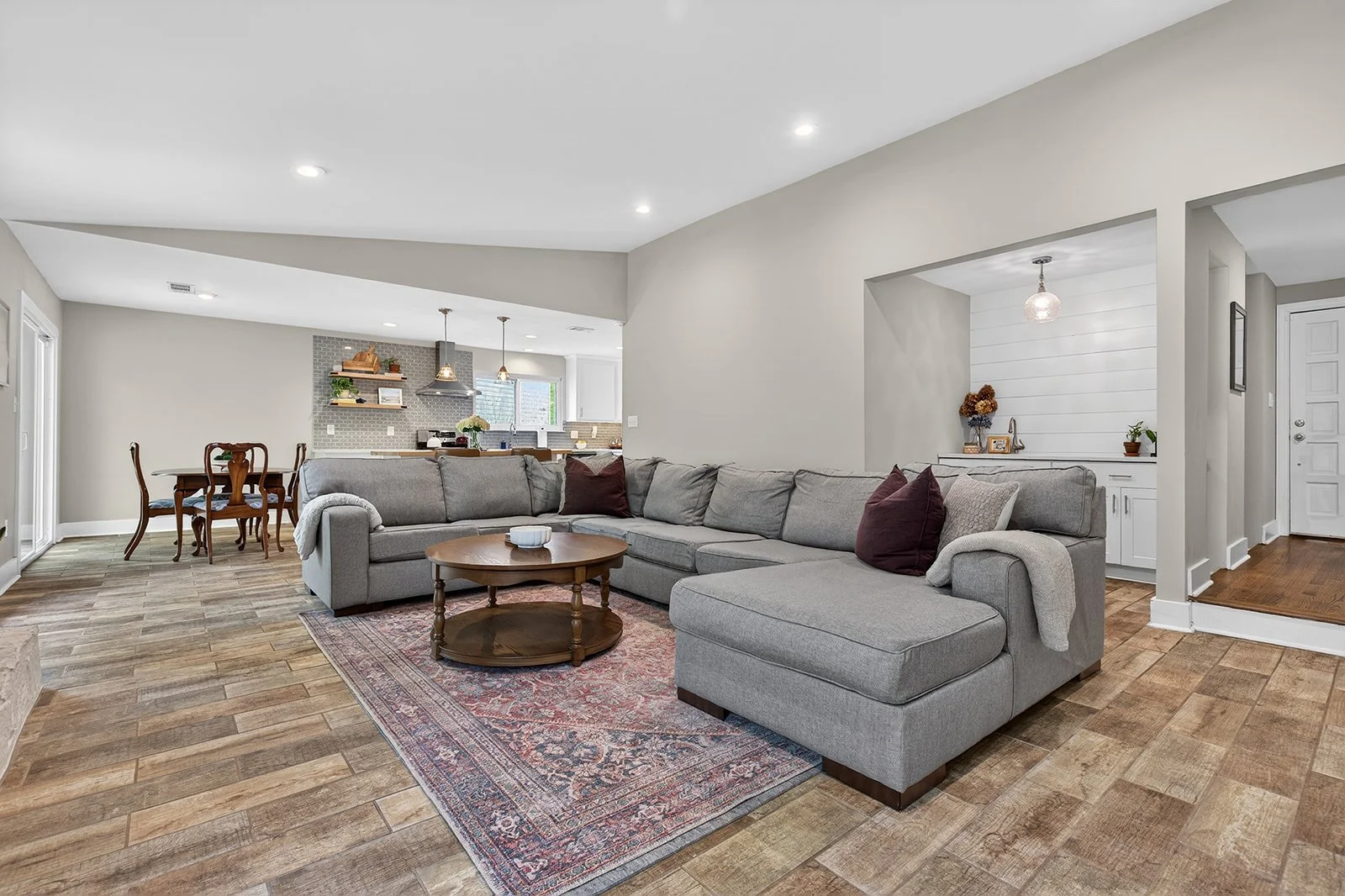 Modern living room with a large gray sectional sofa, a round wooden coffee table, and a patterned area rug. In the background, an open kitchen features pendant lights and a dining area with wooden chairs. The room has wood-look flooring, neutral wall