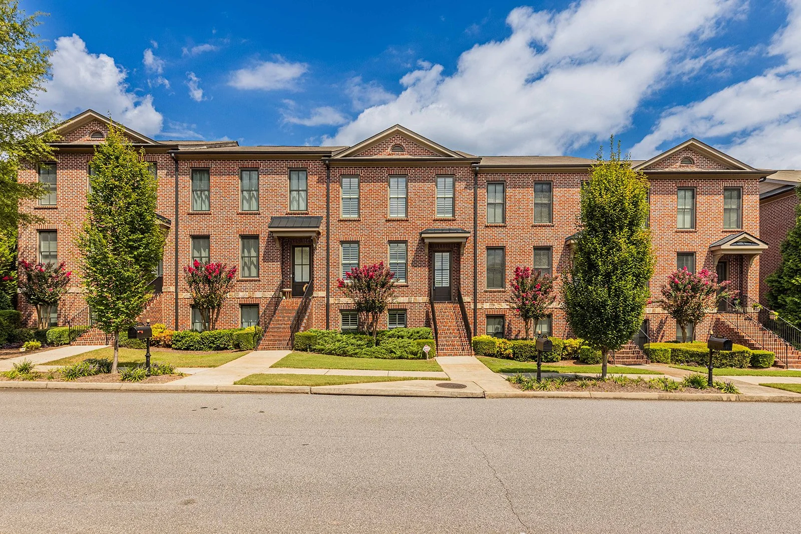 A row of three-story brick apartment buildings with front stairs, green lawns, trees, and flower bushes, under a partly cloudy blue sky.