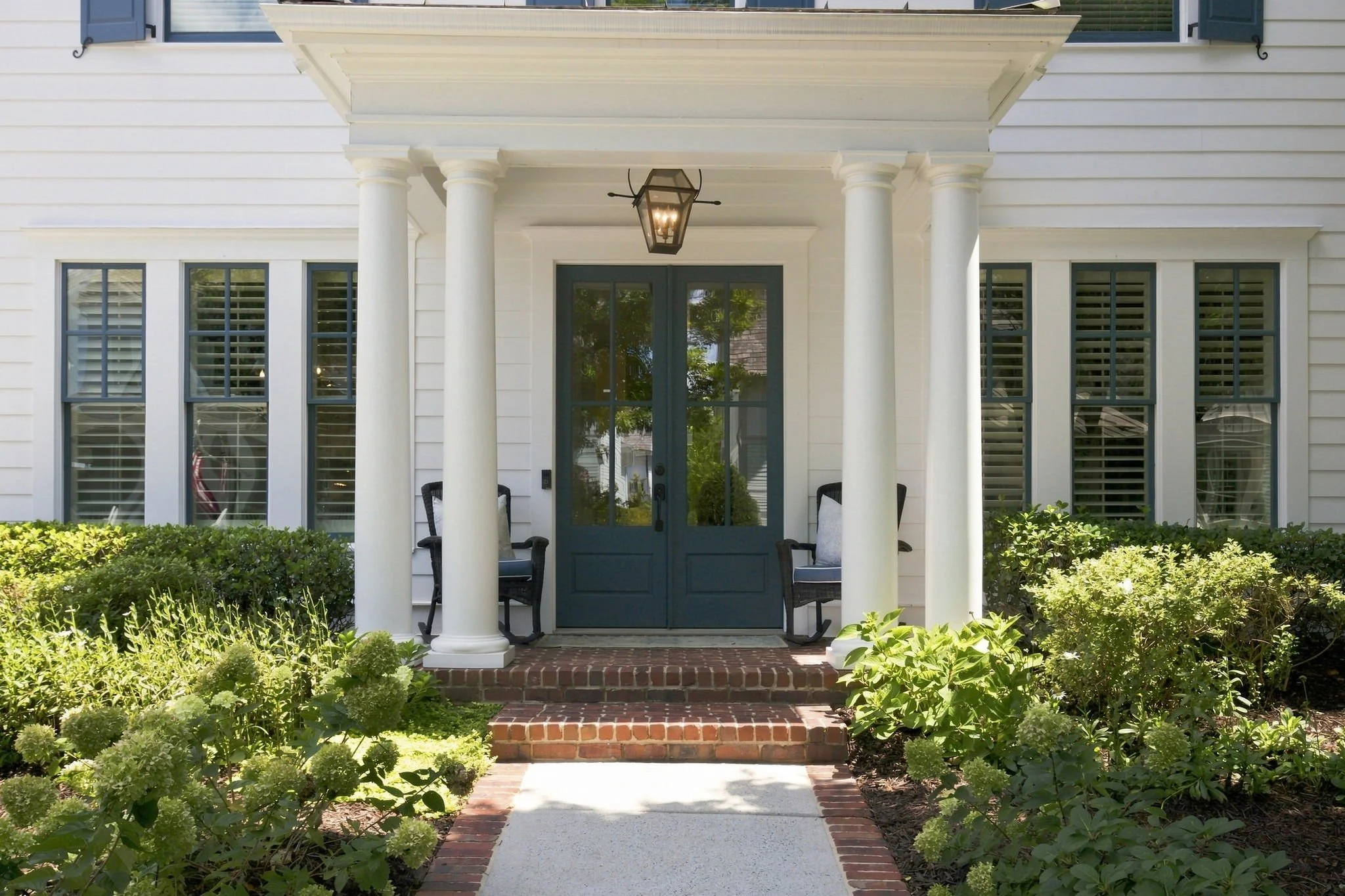 Front porch of a house with white siding, green door, two chairs, and a decorative lantern hanging from the porch ceiling. There is a brick and concrete walkway leading up to the porch and surrounded by lush green bushes.