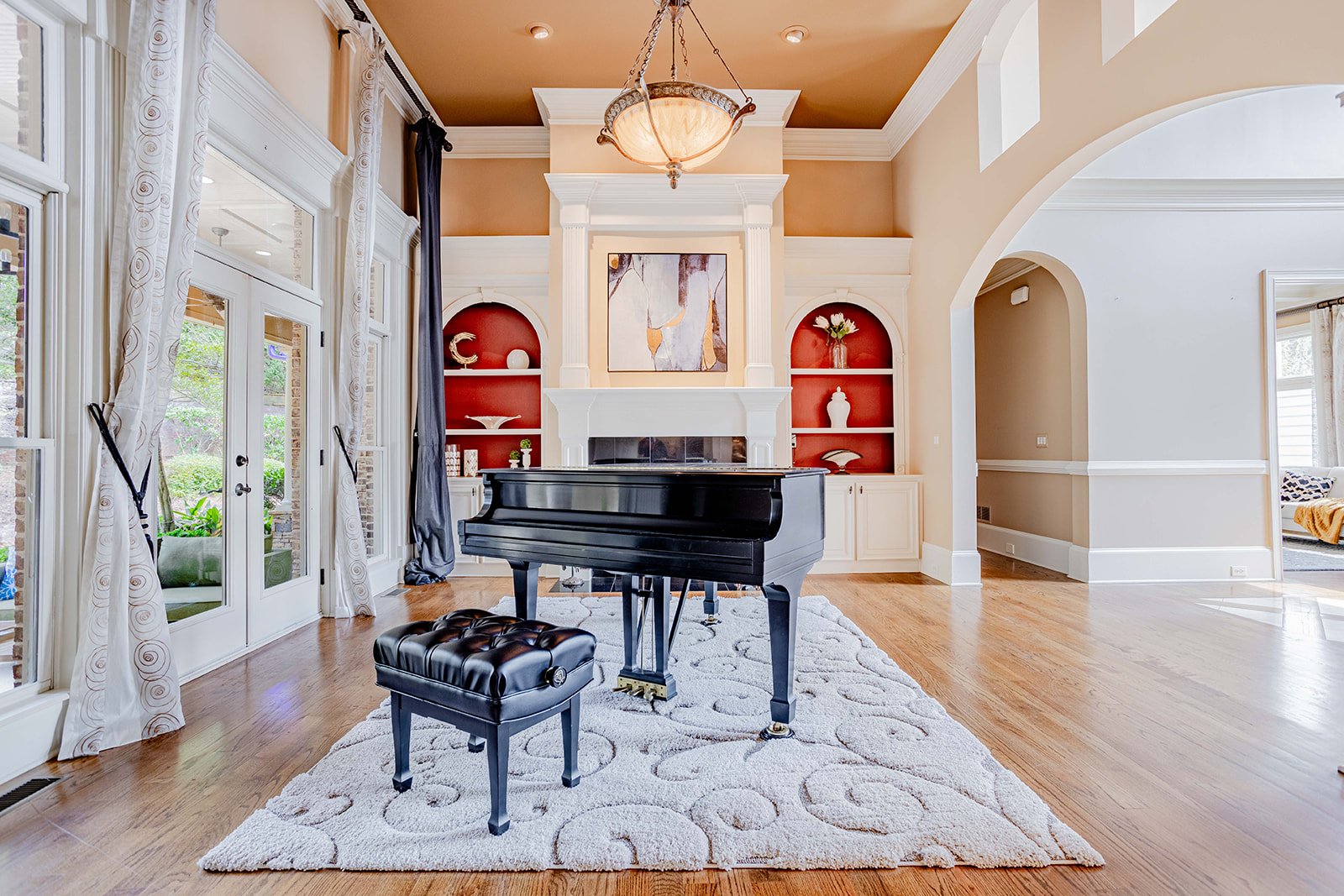 Living room with a black grand piano and matching bench on a cream patterned rug, featuring a fireplace with built-in shelves, a painting above the fireplace, high ceilings, gold ceiling, and large windows with white curtains.