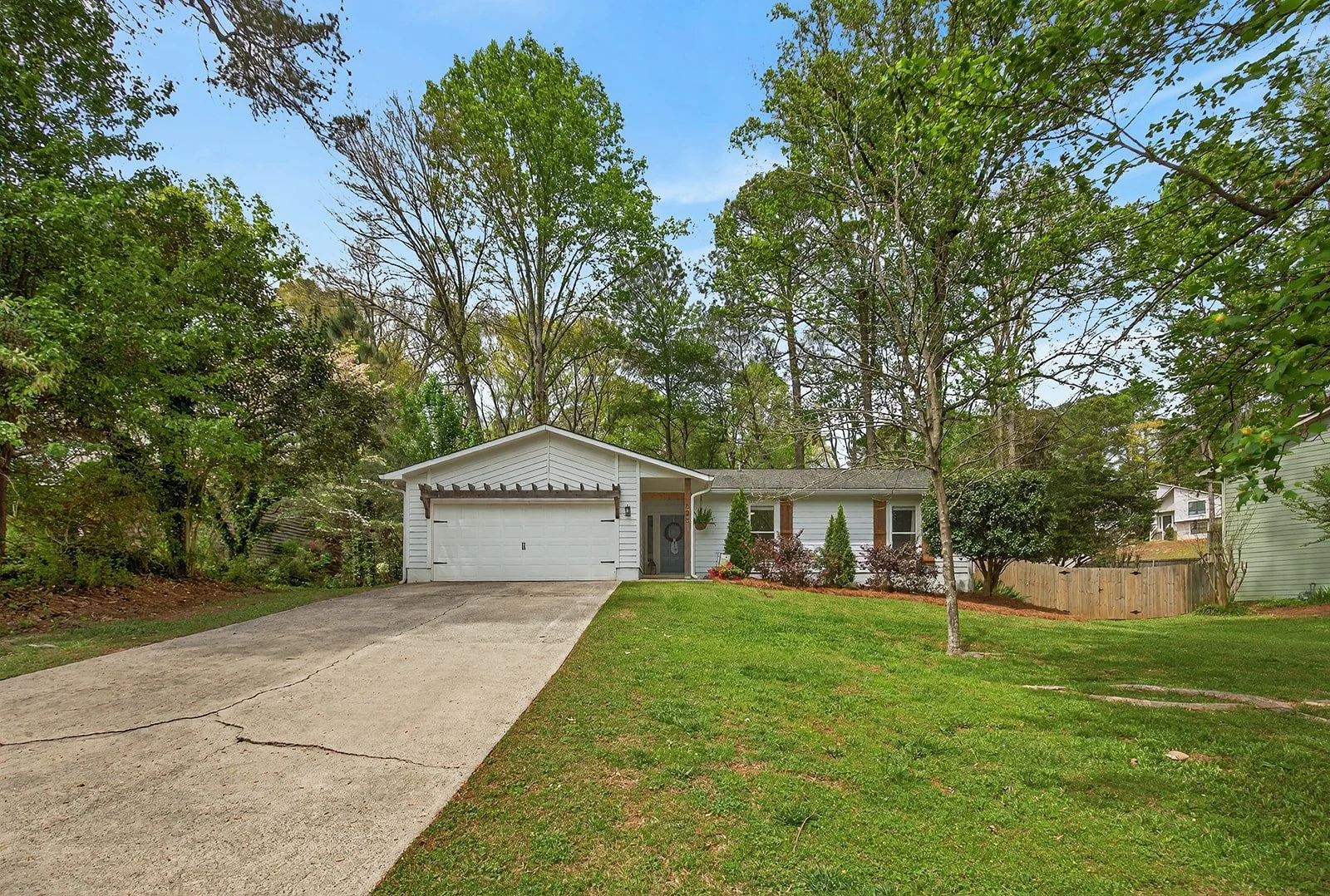 A single-story white house with a gable roof, a two-car garage, and a small front porch area with plants and shrubs. There is a driveway leading to the garage, a well-maintained green lawn, and tall trees surrounding the house under a partly cloudy s