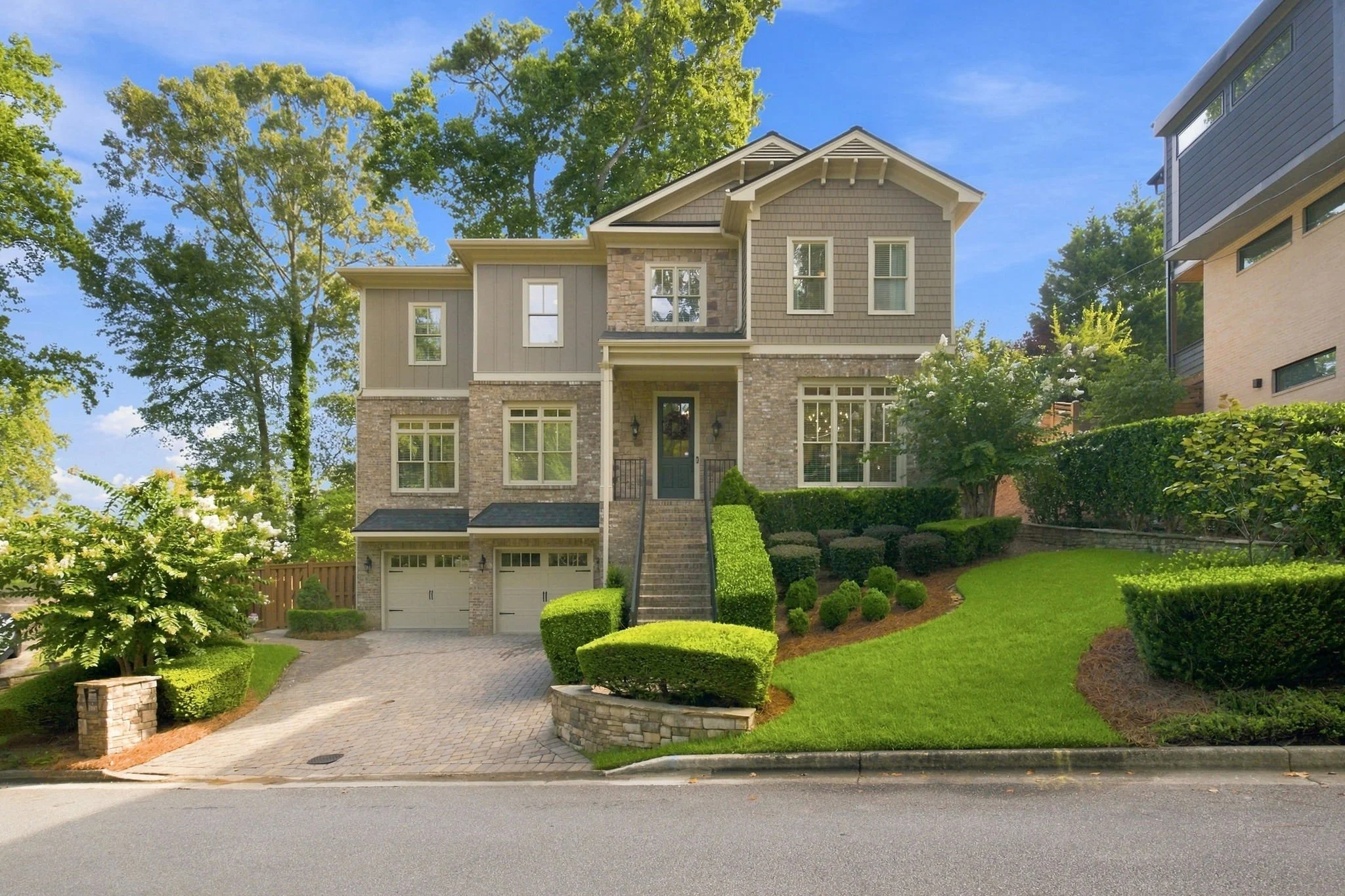 A two-story house with a front yard, driveway, and landscaping. The house has brick and siding exterior with multiple windows and a front door. There are trees and bushes surrounding the property under a blue sky.