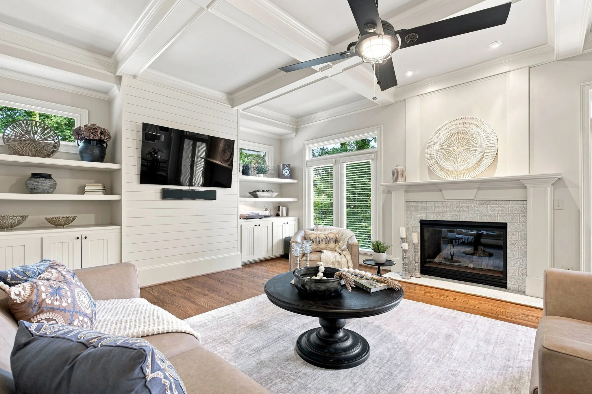 Living room with white walls, large windows, a fireplace, a ceiling fan, built-in shelves decorated with vases and books, a black coffee table, beige sofas with patterned pillows, and hardwood flooring.