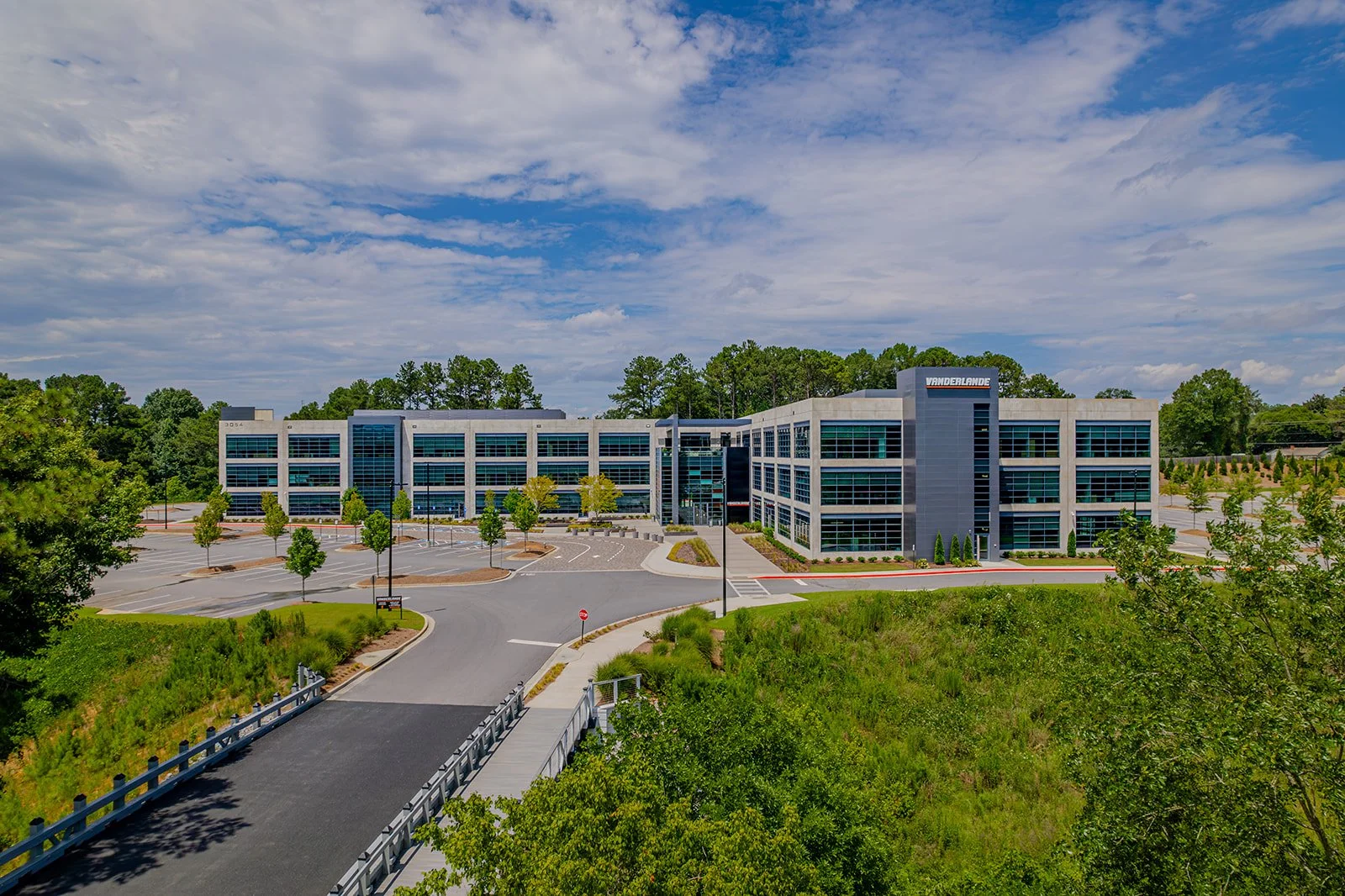 A modern multi-story office building with large glass windows, surrounded by an empty parking lot with marked spaces and a landscaped area with small trees and shrubs.