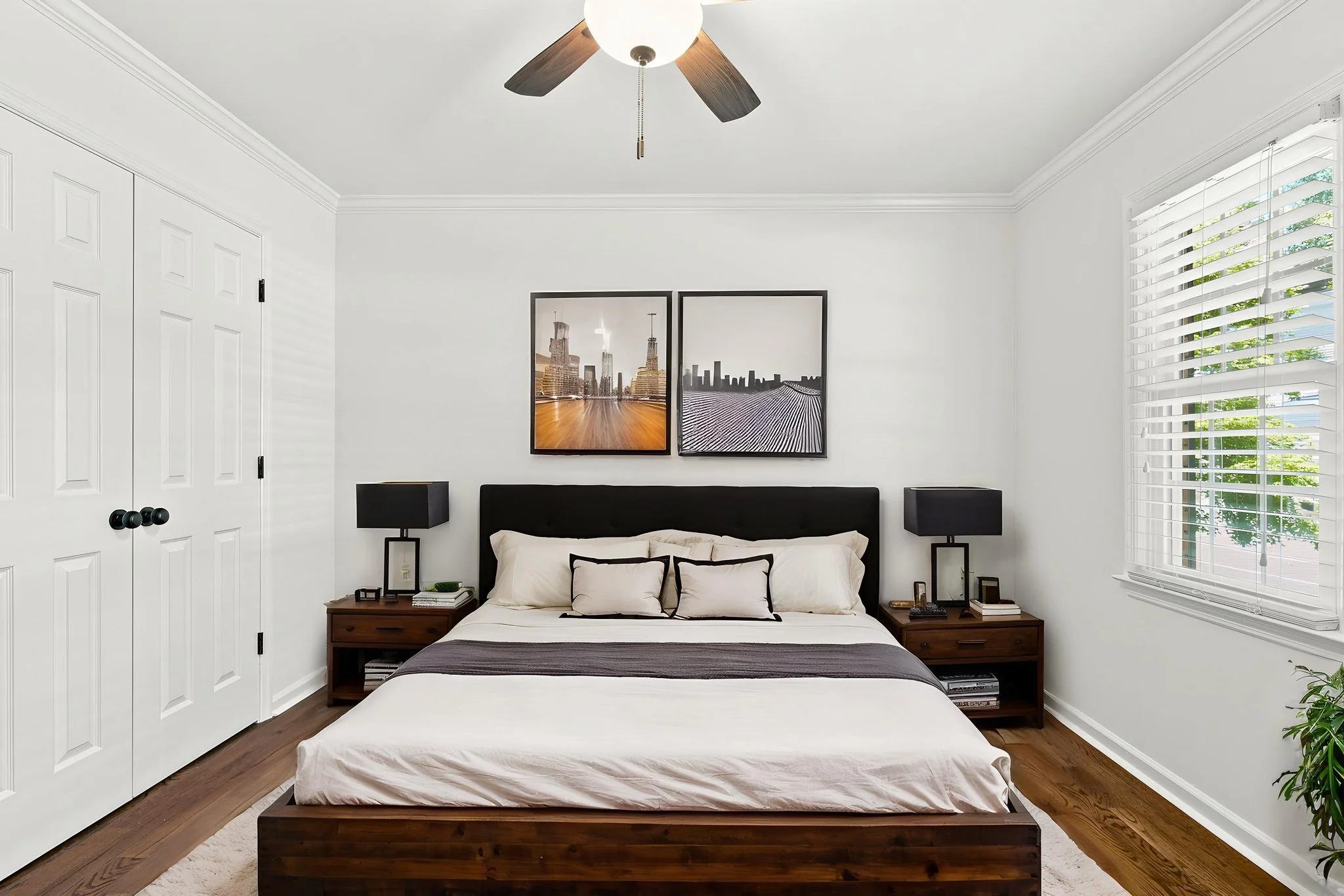 A bedroom with a white bed, black headboard, and matching black nightstands on each side. There are two black lamps on the nightstands, two framed pictures on the wall above the bed, a window with white blinds on the right, and a closet with white double doors on the left. The room has white walls, crown molding, and hardwood flooring.