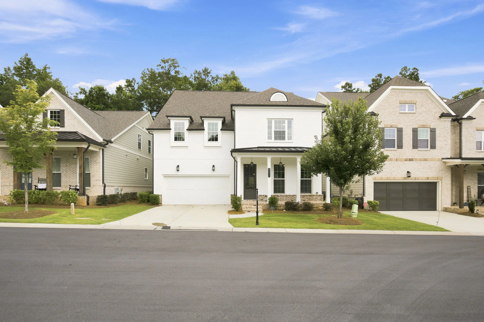 Modern two-story suburban house with white and beige brick exterior, gray shingle roof, front porch, two small trees, green lawn, sidewalk, and street.