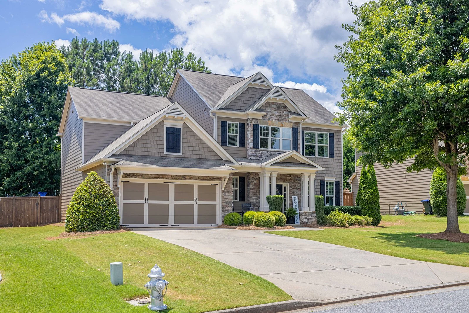 A two-story suburban house with gray siding, black shutters, a stone facade, and a two-car garage, surrounded by green grass, trees, and shrubs under a partly cloudy sky.