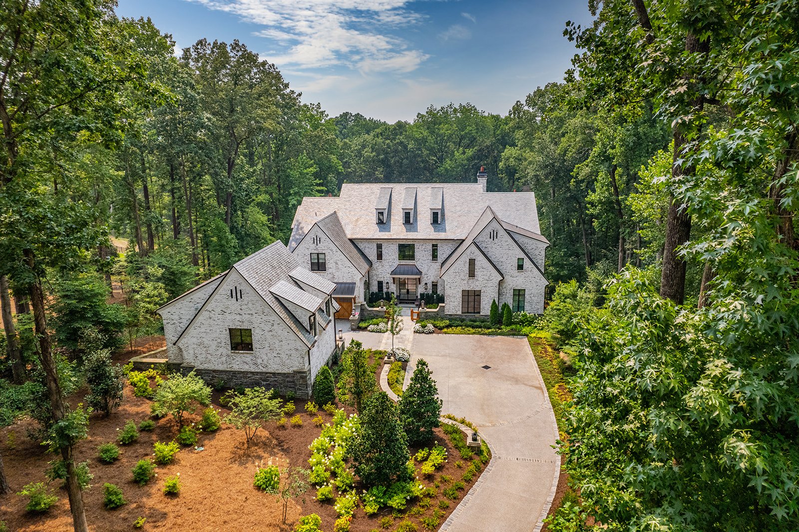 A large white brick house with multiple gables and dormer windows, surrounded by lush green trees and a landscaped garden with small bushes and plants, and a wide driveway leading to the front entrance.