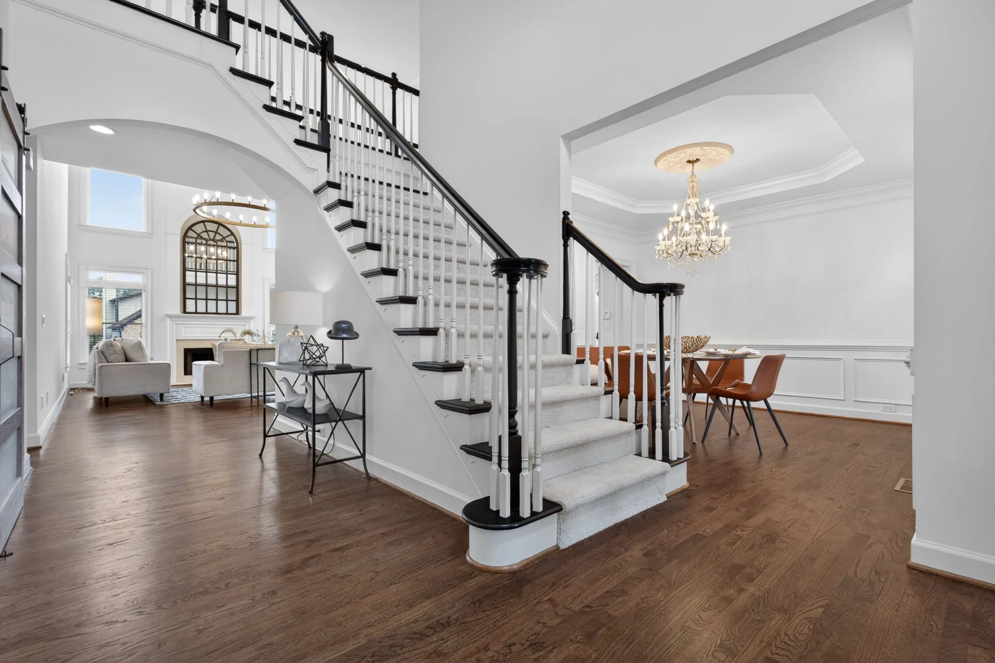Interior of a modern home featuring a staircase with black and white railings, hardwood floors, a chandelier, a living room with sofas and a fireplace, and a dining area with a table and orange chairs.