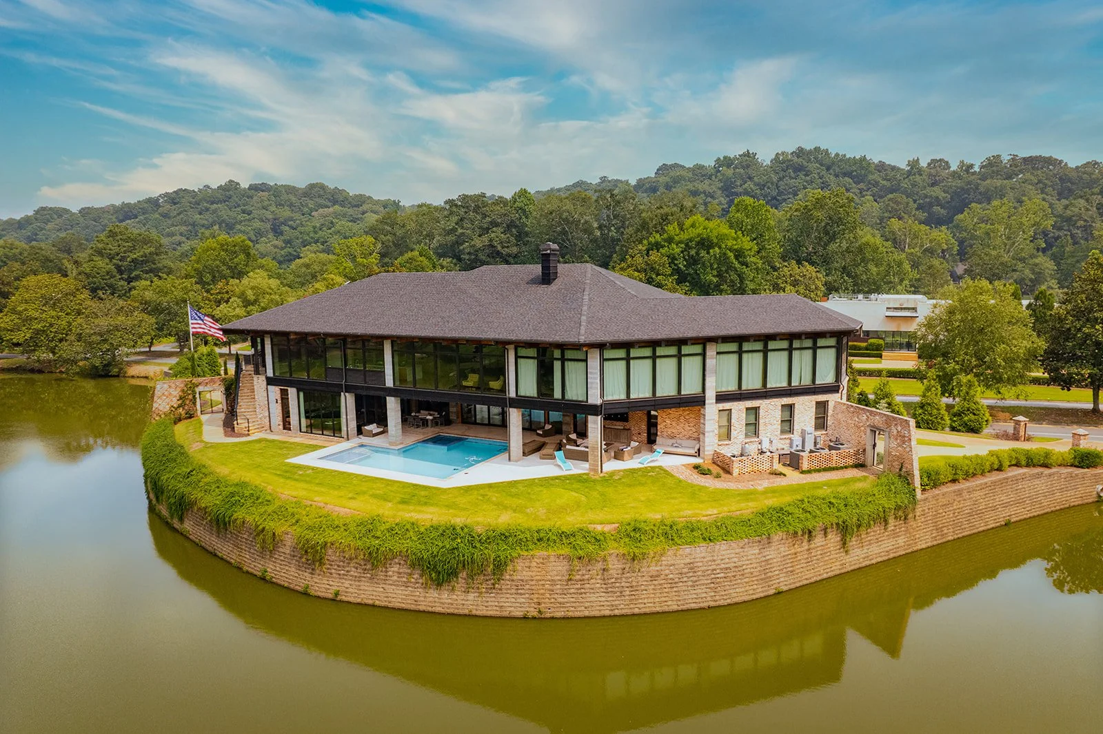 A large modern house with glass walls and a pitched roof, situated on a small island surrounded by water, with an outdoor pool and lush green landscape.
