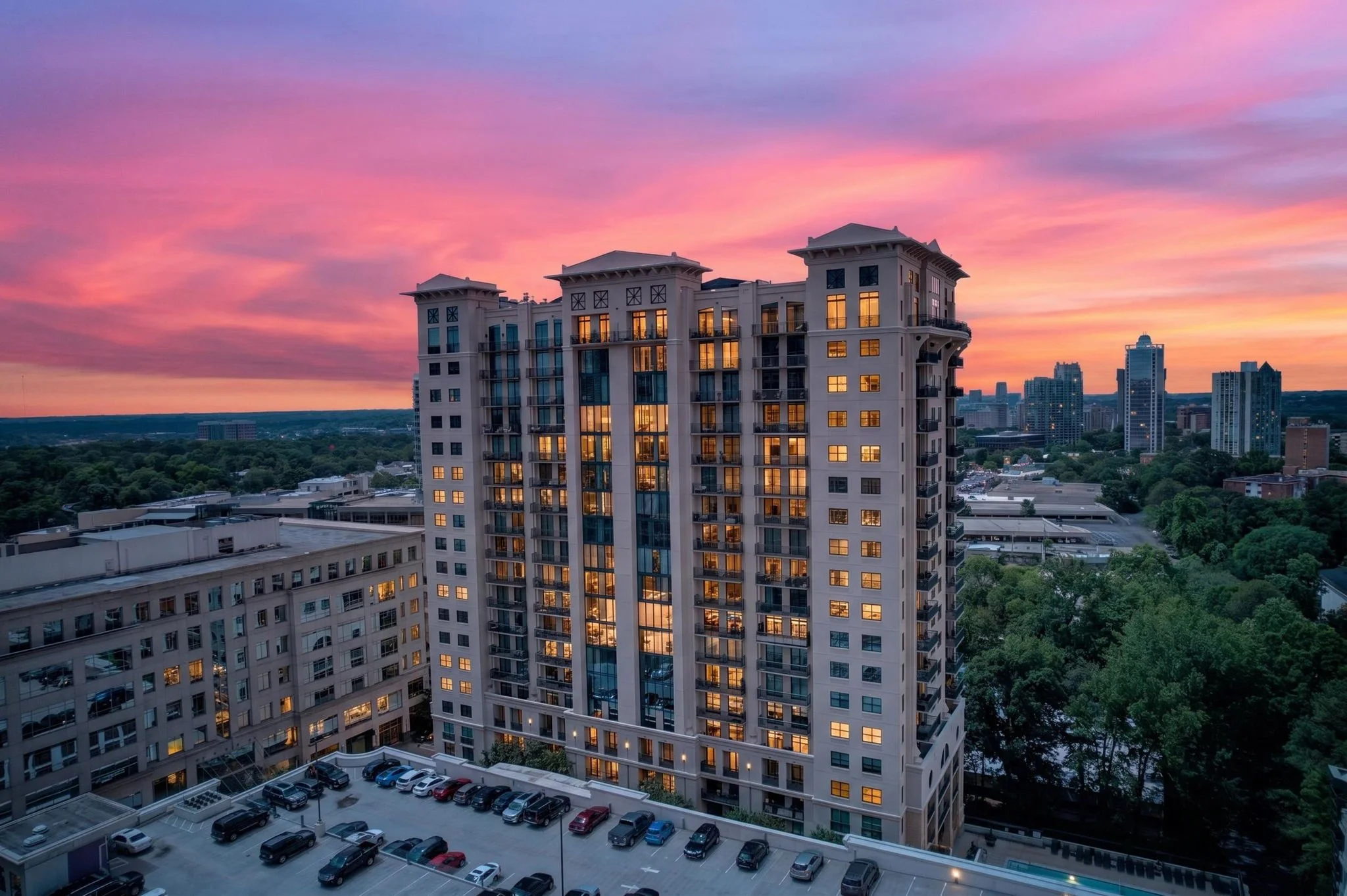 High-rise apartment building during sunset with illuminated windows, city skyline in the background, and a parking lot filled with cars in the foreground.